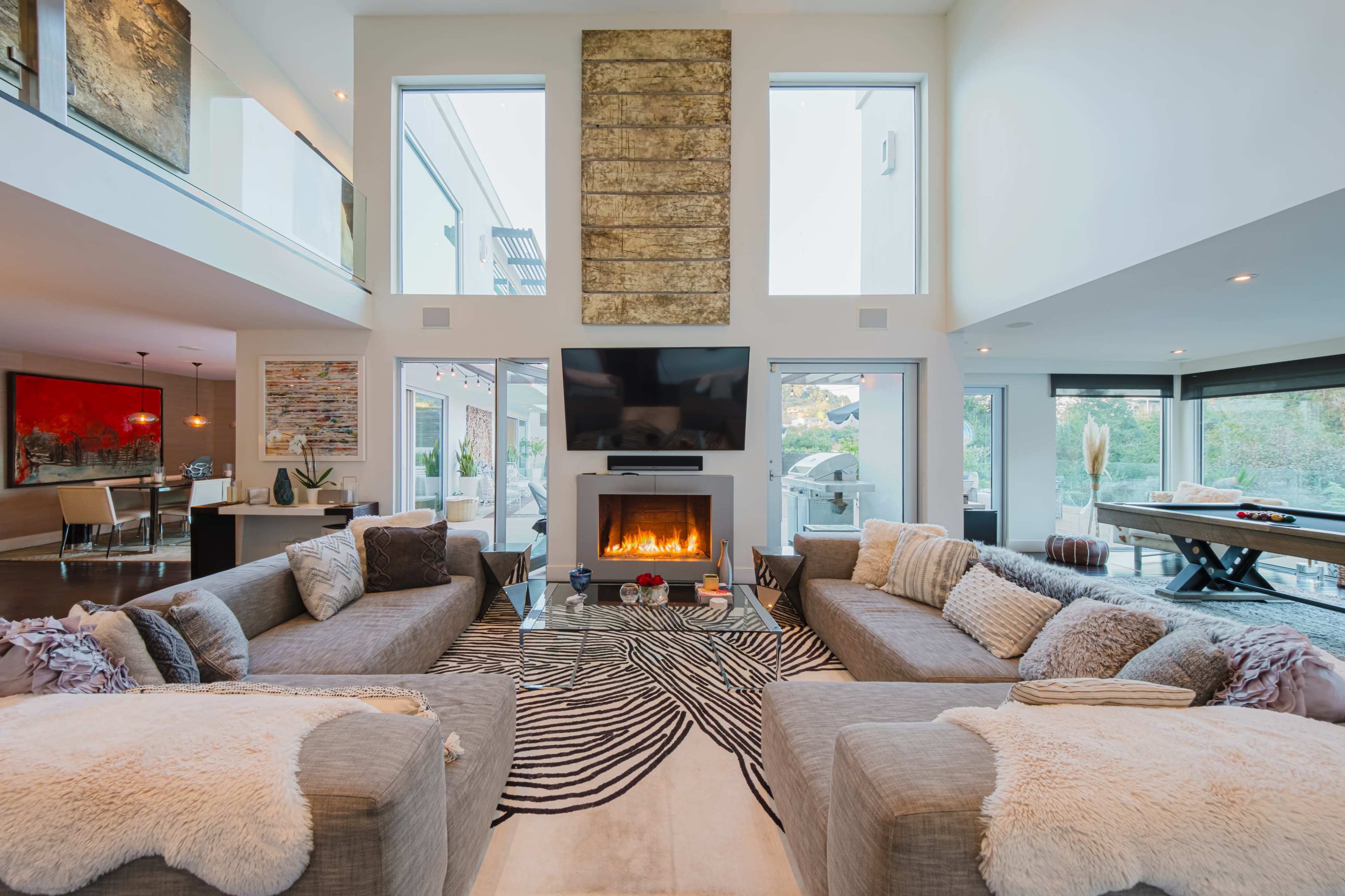 The image shows a modern living room with two large sectional sofas facing a central fireplace, flanked by large windows and an open dining area in the background.