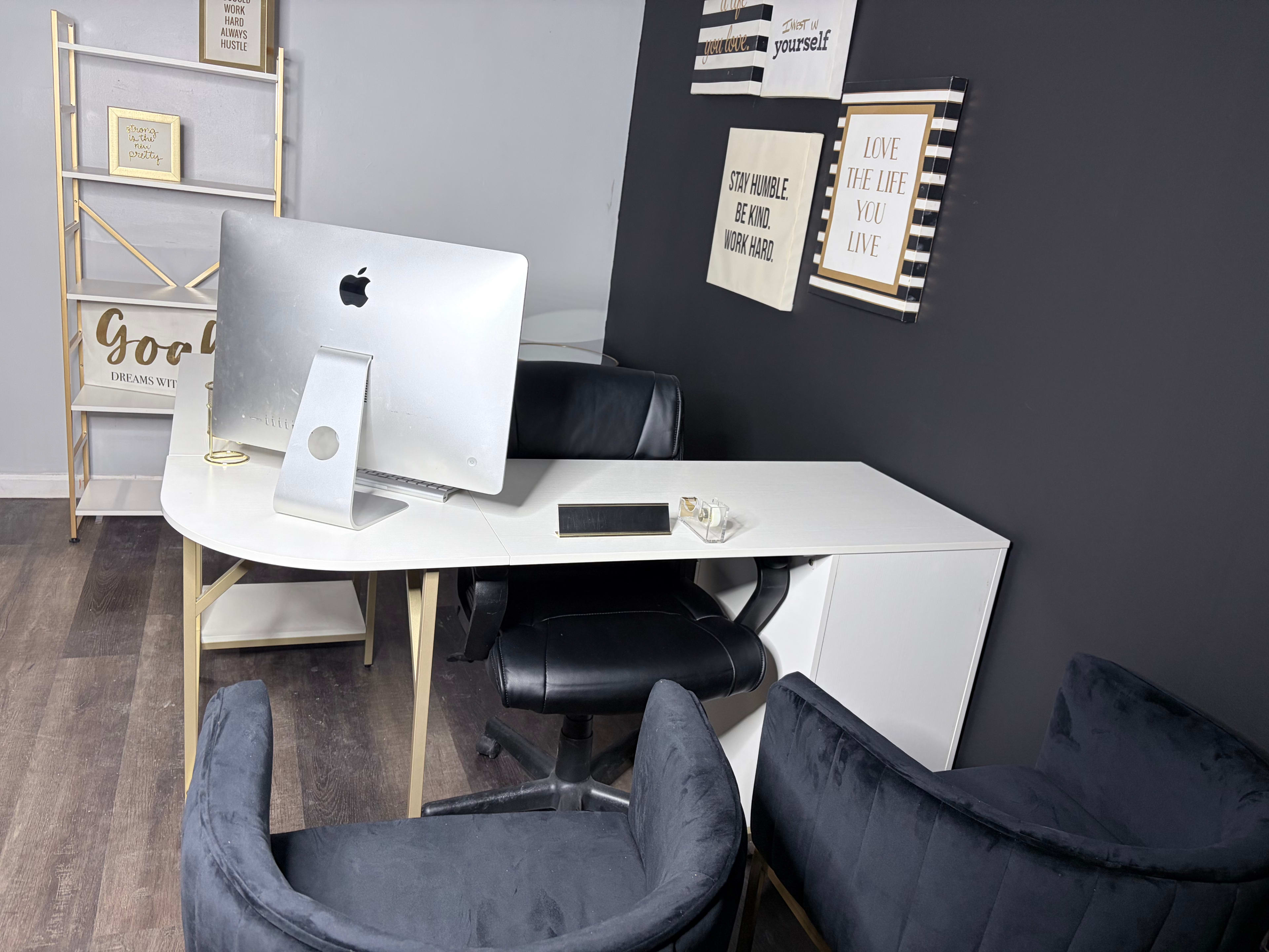 The image shows a modern office space featuring a white desk with a computer, two black chairs, and blue upholstered armchairs against a dark wall decorated with framed quotes.