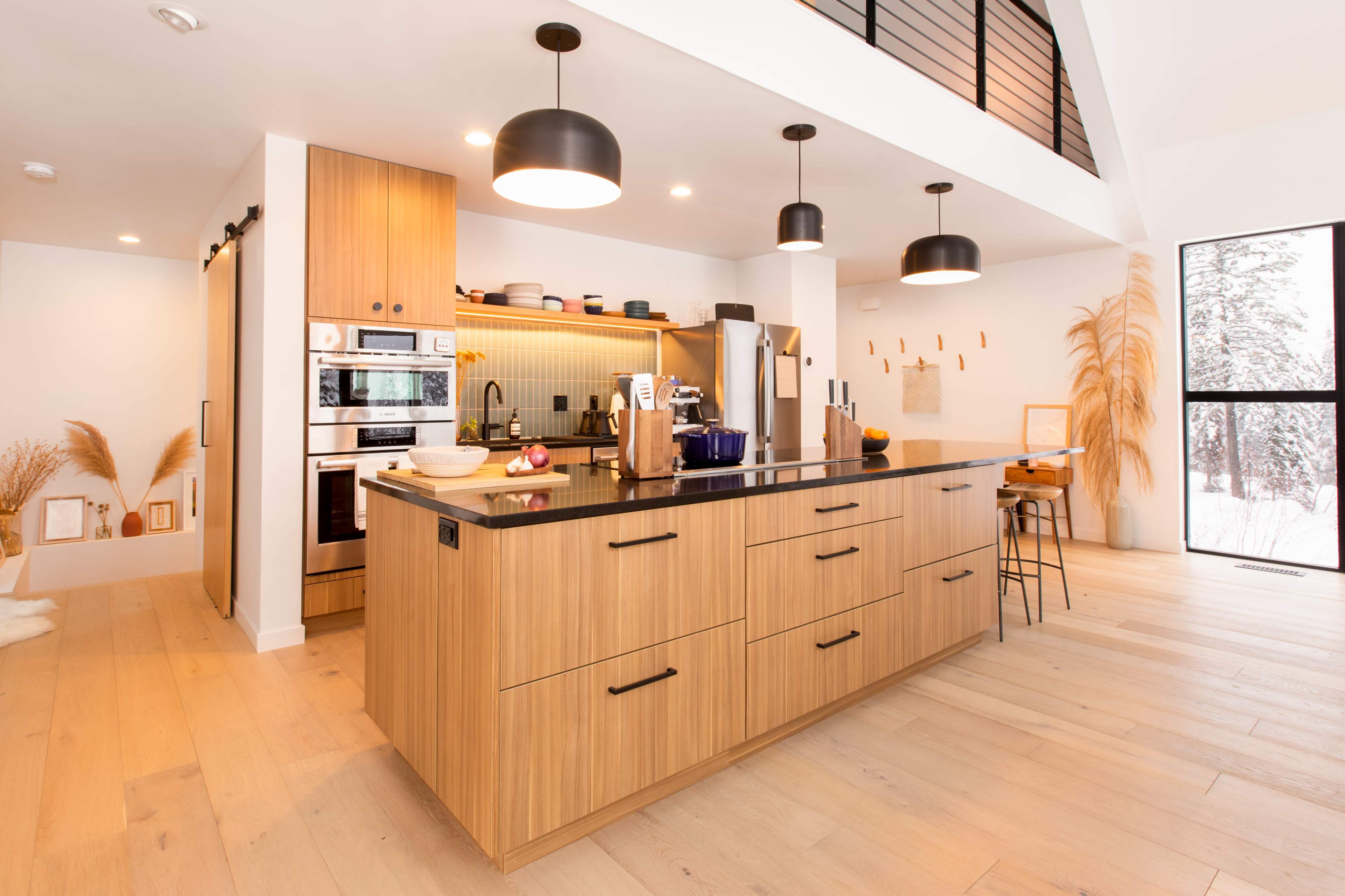 A modern kitchen features a central island with wooden cabinetry, integrated appliances, and barstool seating, surrounded by large windows showcasing a snowy outdoor view.