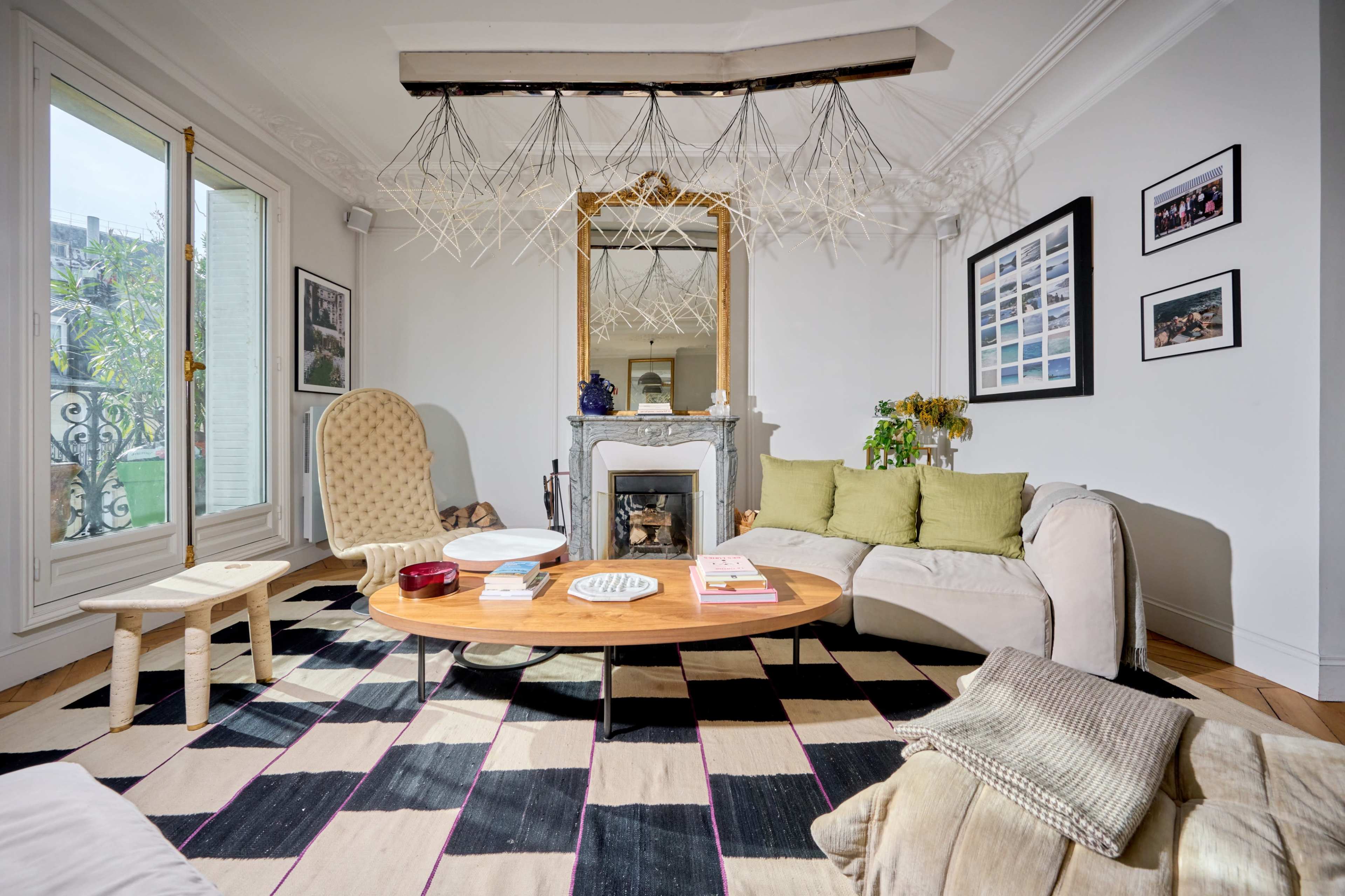 The living room features a round wooden coffee table, a beige sofa with green cushions, and a patterned black and white rug, with large windows and decorative wall art.