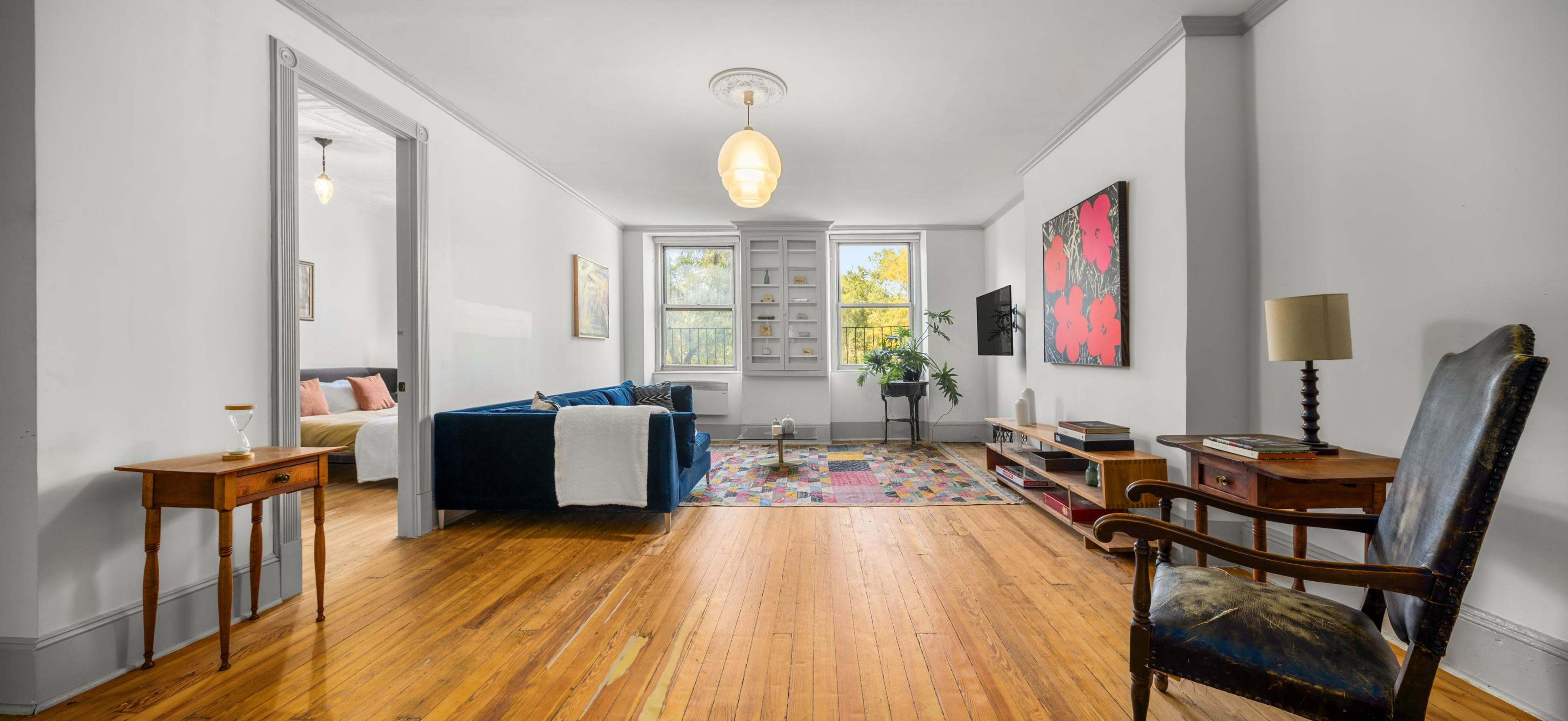 A well-lit living room features hardwood floors, a blue sofa, a colorful rug, and a mix of furniture, including a wooden chair and a low bookshelf.