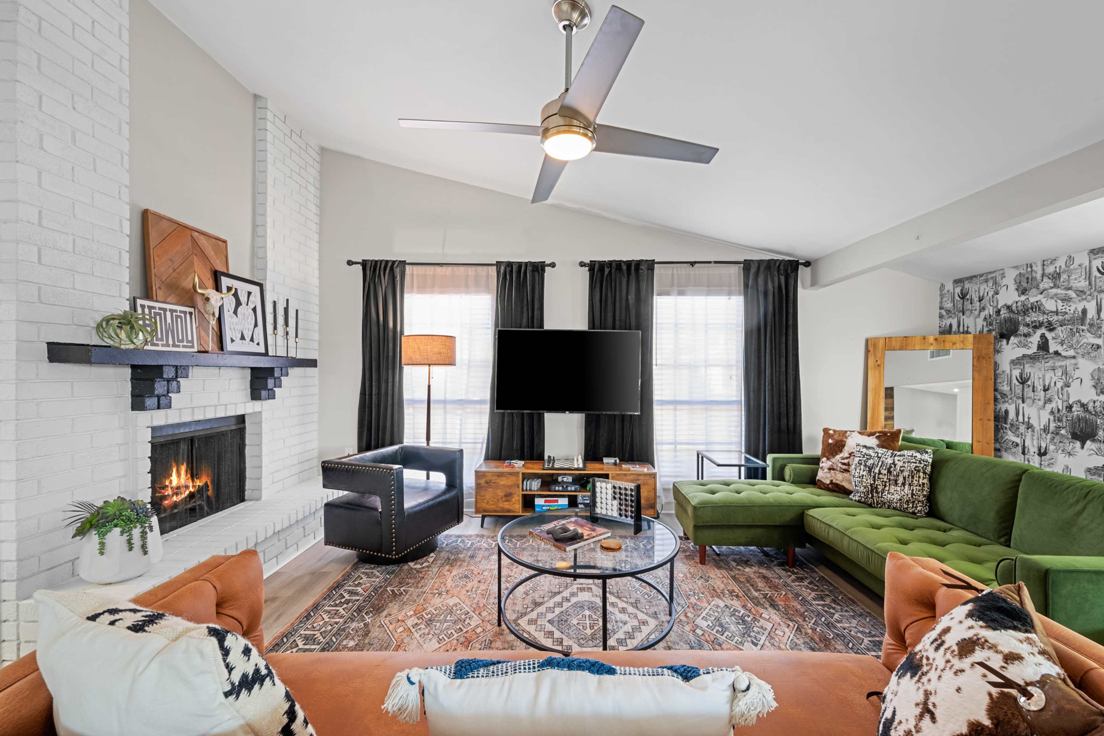 A modern living room features a green sectional sofa, an orange couch, a circular coffee table, and a mounted television above a fireplace, with large windows dressed in black curtains.