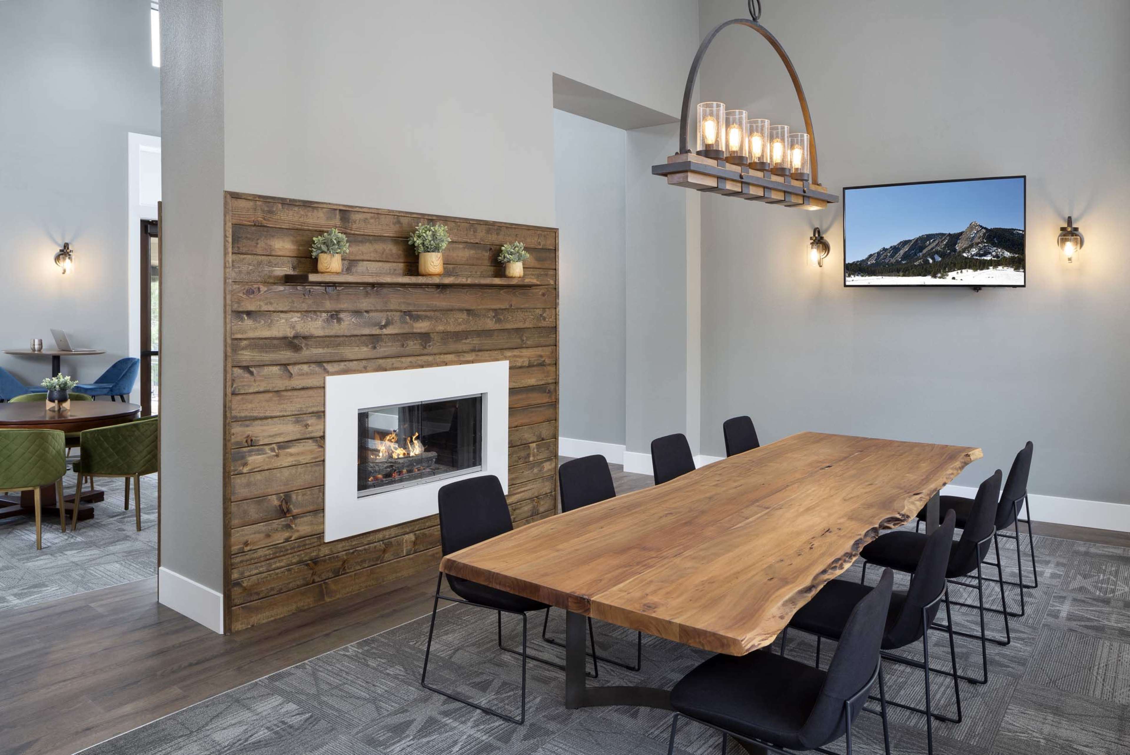 A large wooden table with black chairs is positioned in a well-lit room featuring a modern fireplace and a mounted television on the wall.