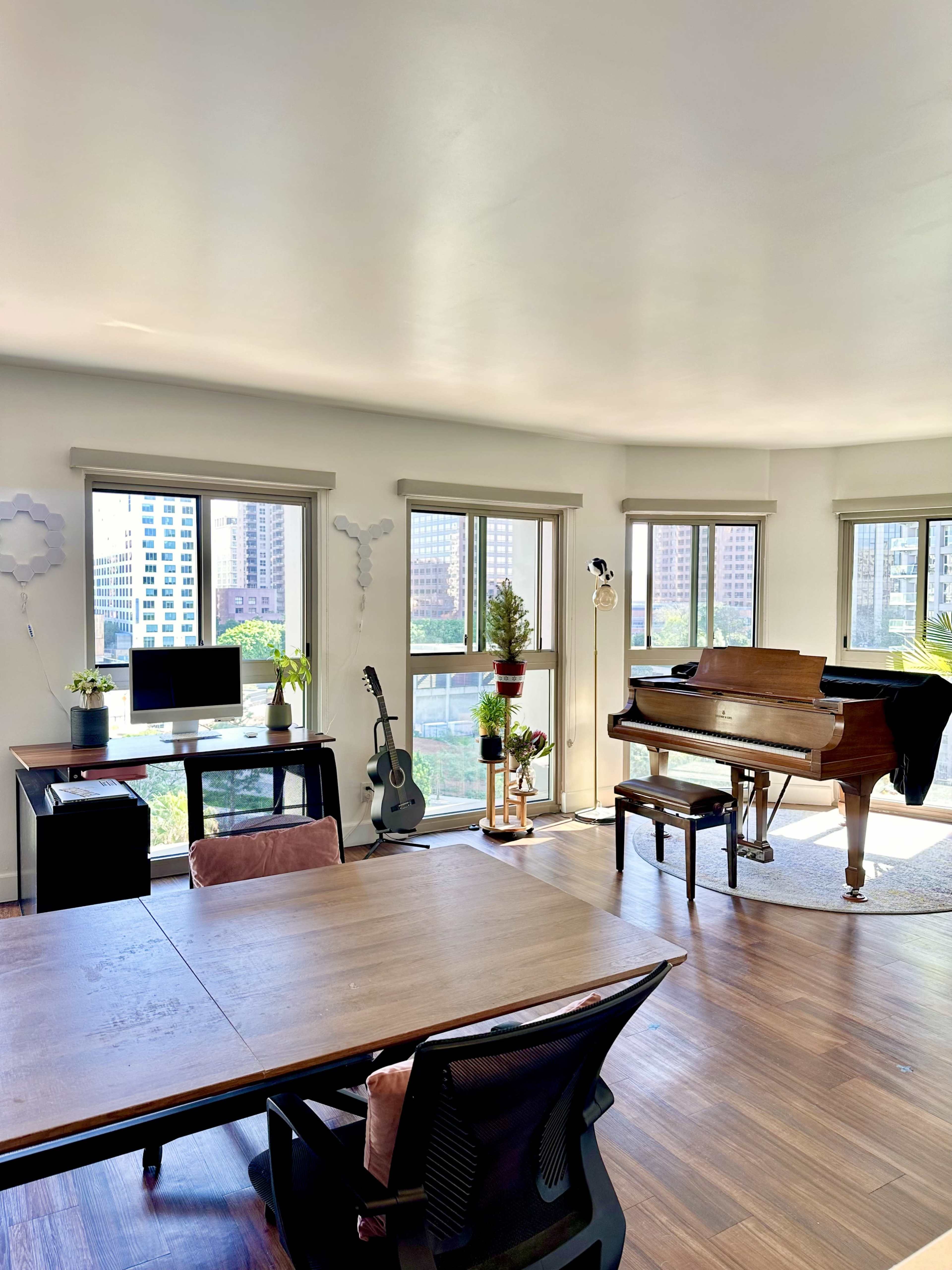 A bright, modern room features a piano, a desk with a computer, and large windows with city views.