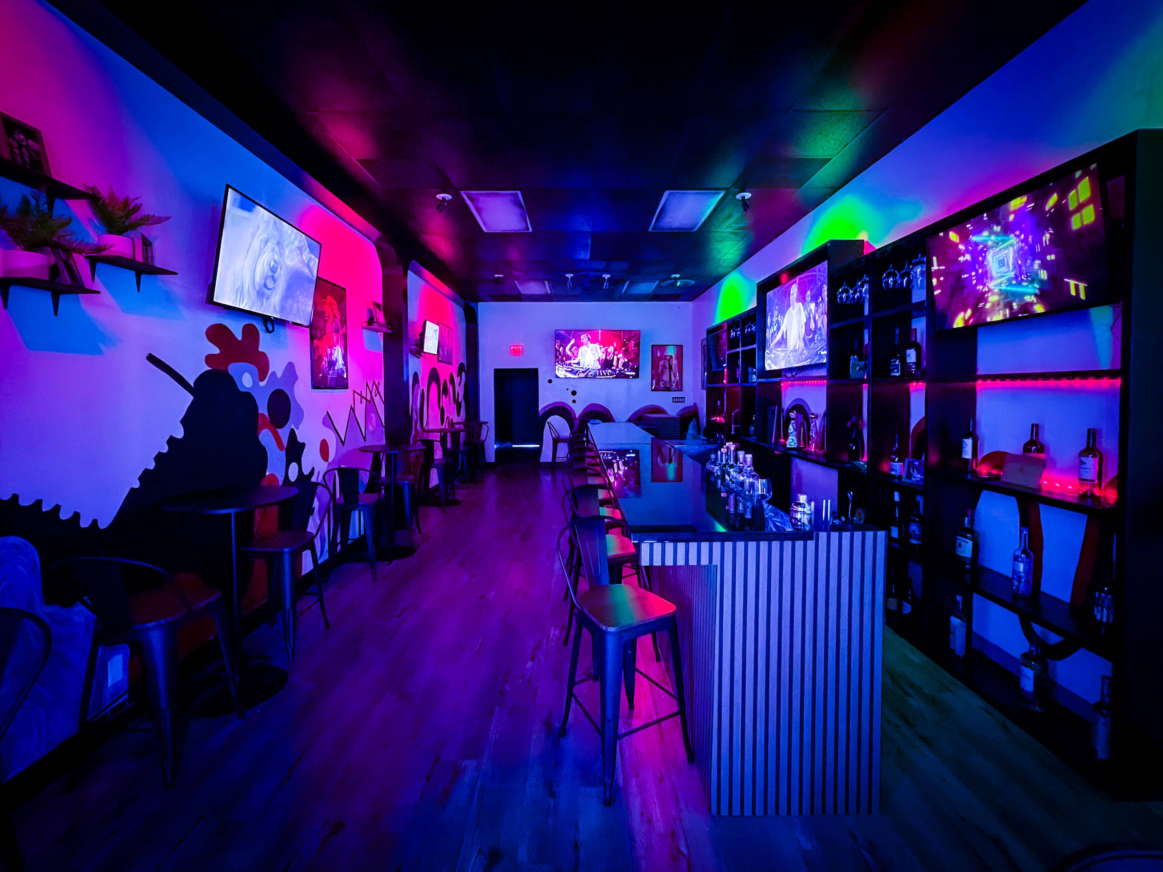A dimly lit bar interior features colorful lighting, multiple TVs, and seating arranged around a central counter.