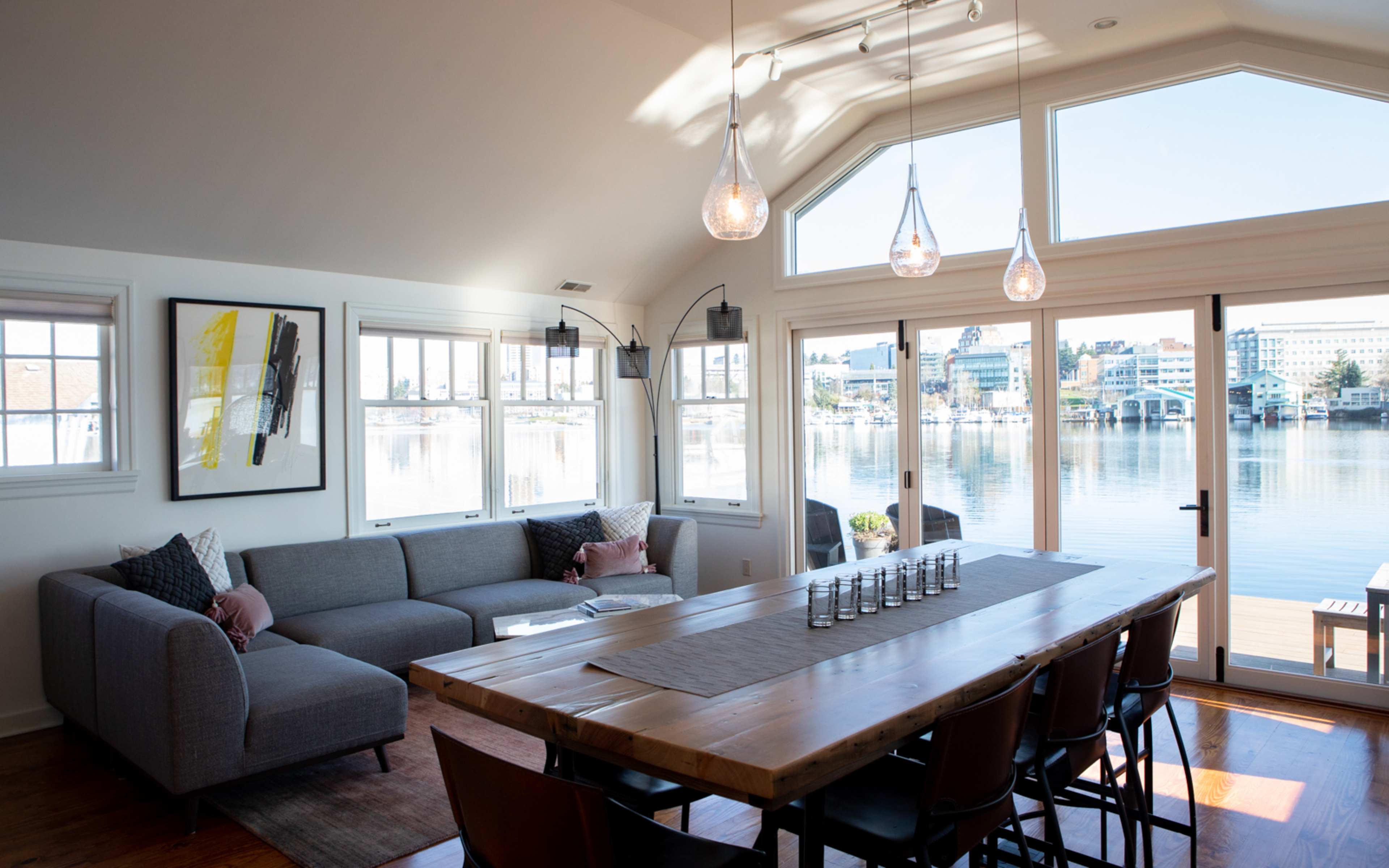 A modern living space features a large gray sofa, a wooden dining table with chairs, and expansive windows overlooking a body of water.