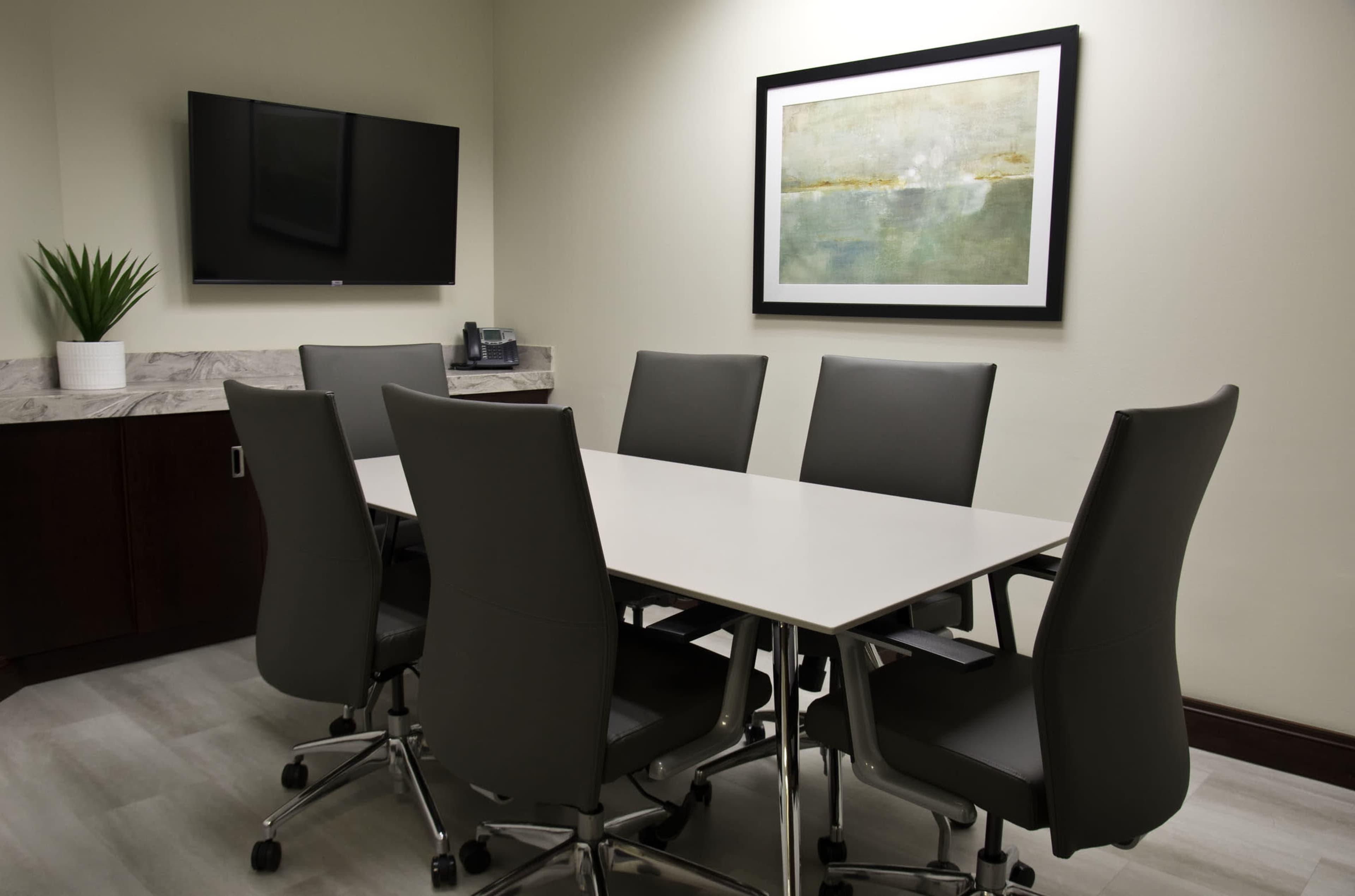 A modern conference room features a rectangular table surrounded by six ergonomic chairs, with a television mounted on the wall and a piece of abstract art hanging above.