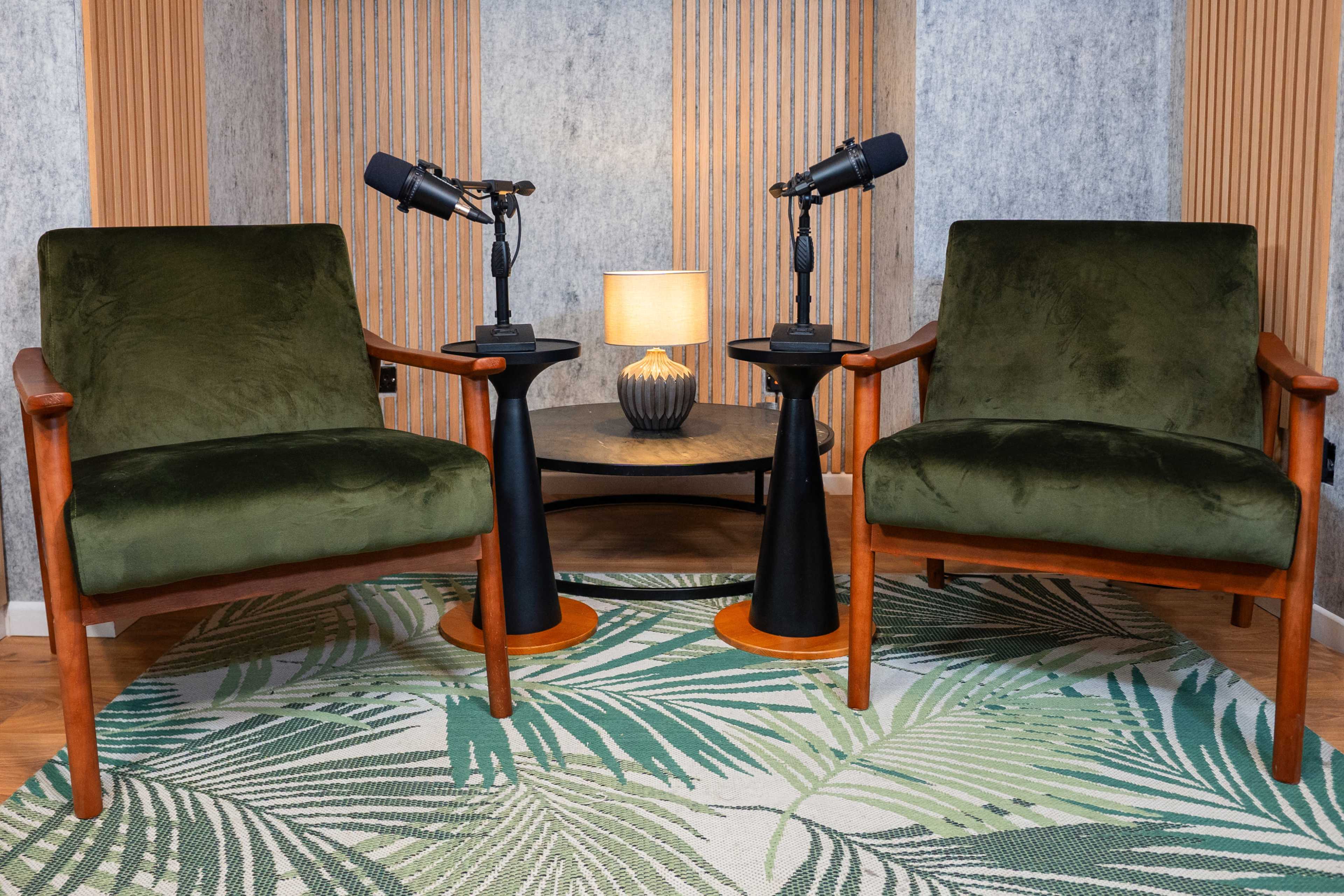 Two green upholstered chairs facing each other, positioned on a palm leaf-patterned rug, with a small table in between holding a lamp and two microphones.