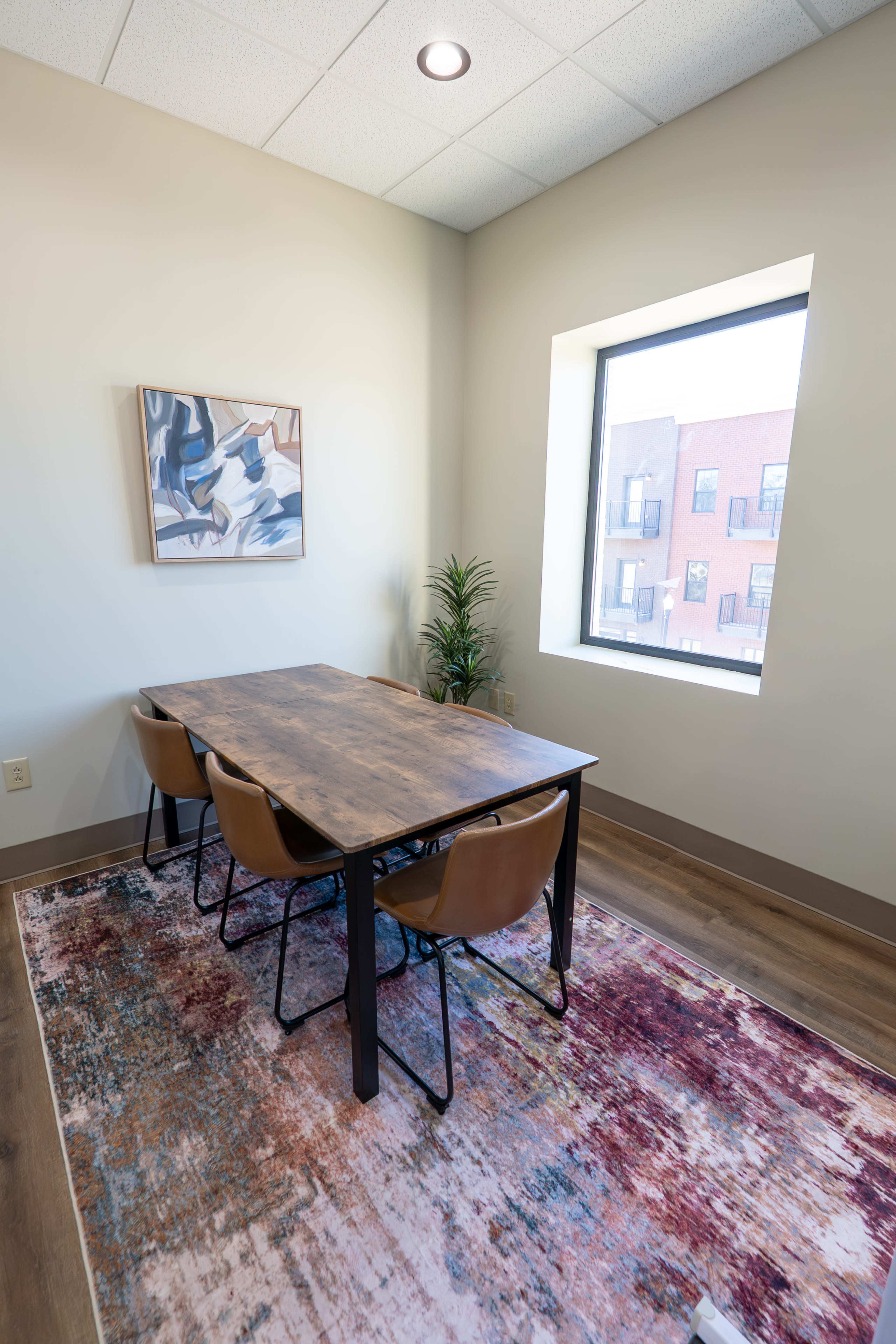 A small meeting room features a wooden table with three brown chairs, a decorative plant, and a window overlooking a building.
