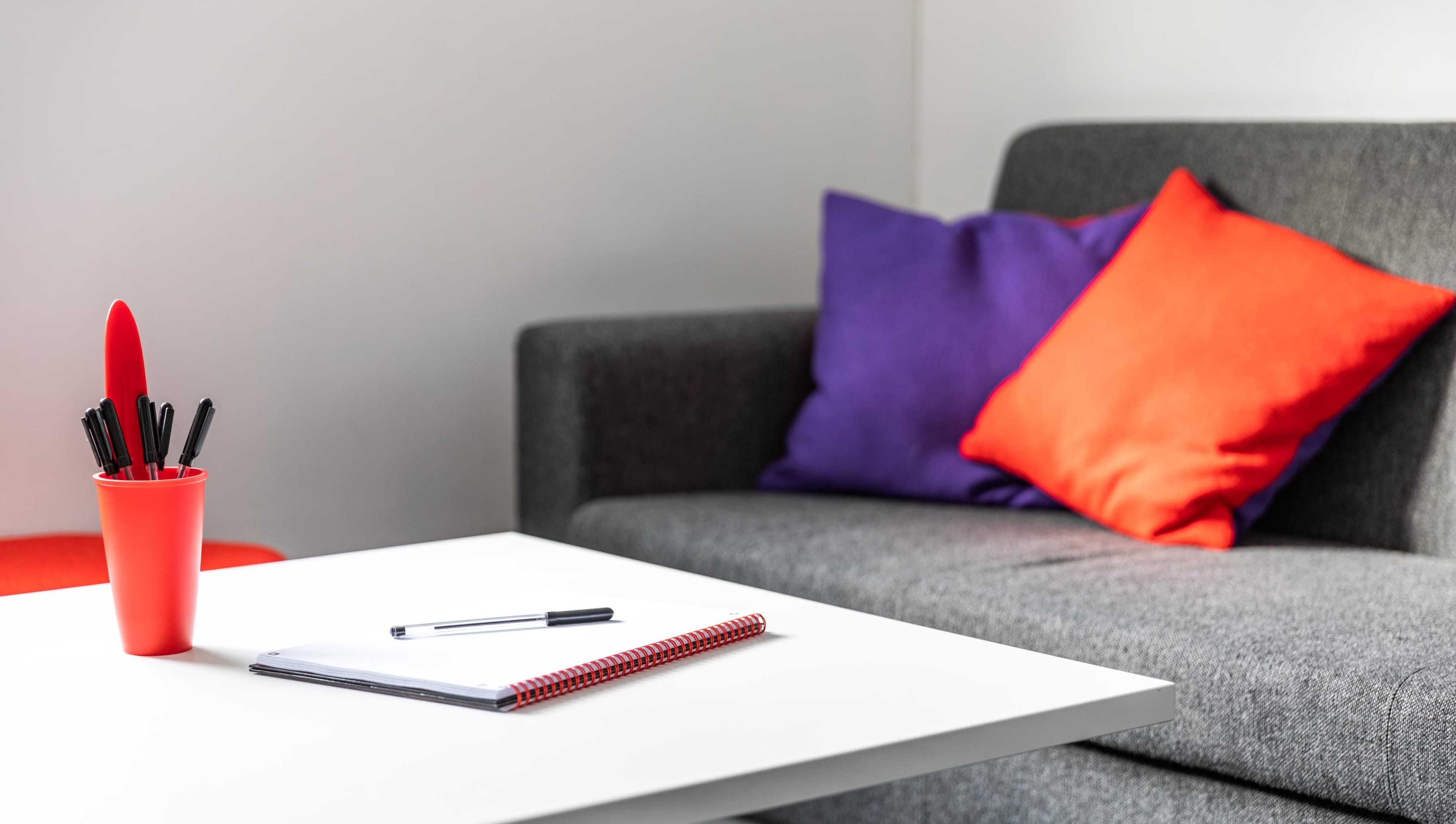A red pen holder filled with pens sits on a white table next to a notebook, while a gray couch with orange and purple cushions is visible in the background.
