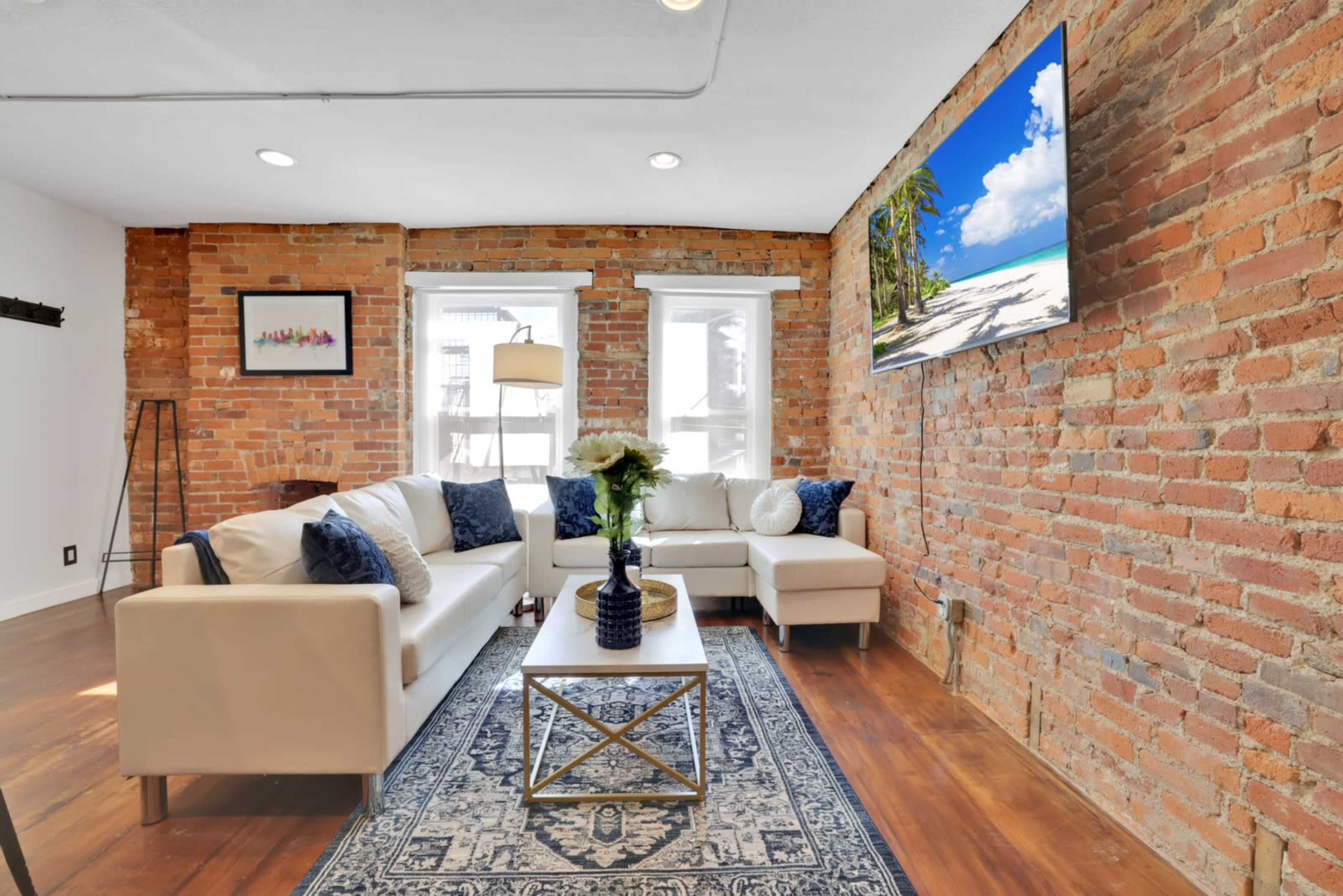 A living room with exposed brick walls, a white sofa, a coffee table, and a television mounted on the wall, featuring large windows that allow natural light.