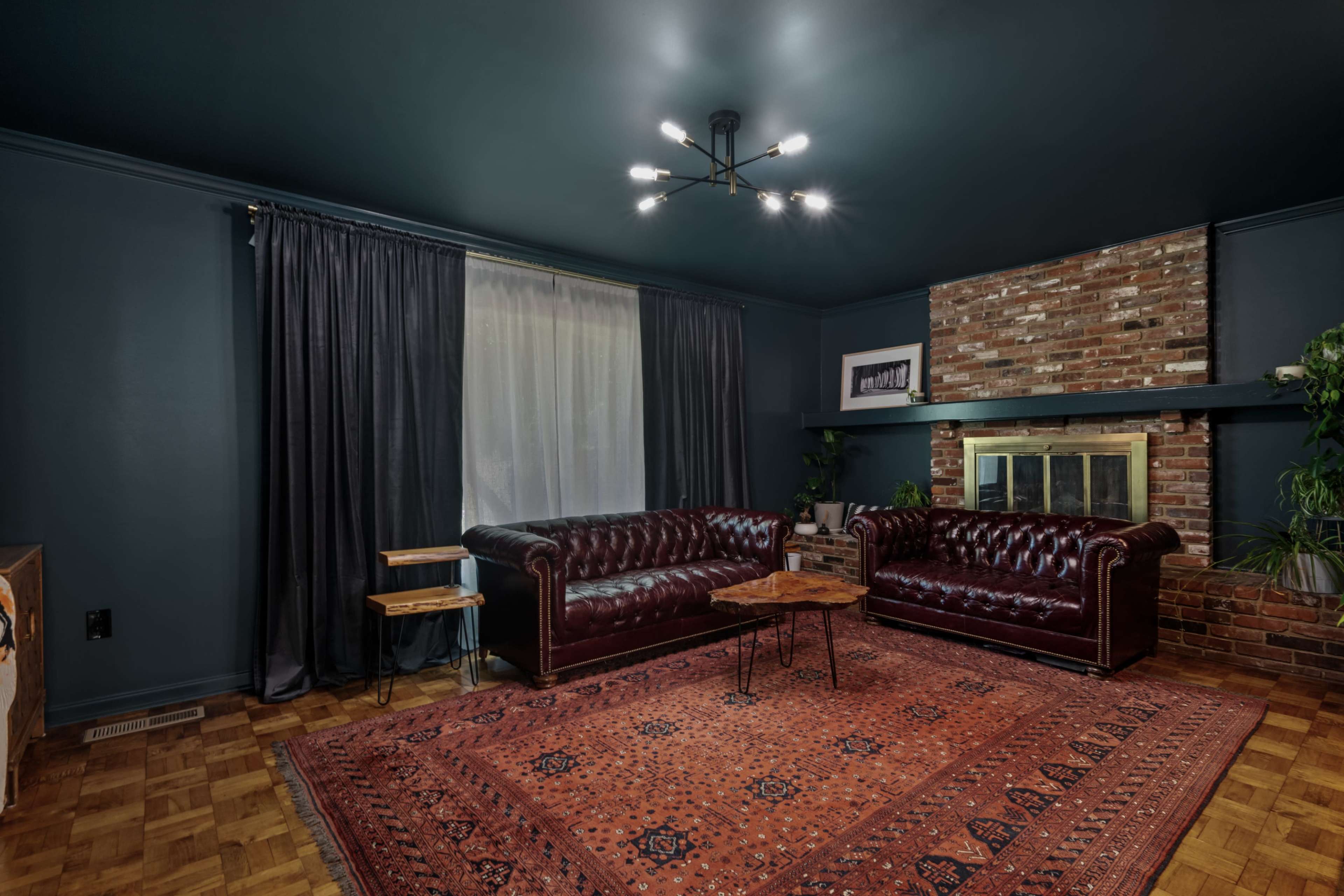 A living room with dark blue walls, a brick fireplace, two deep red leather sofas, a small wooden table, and a large area rug.
