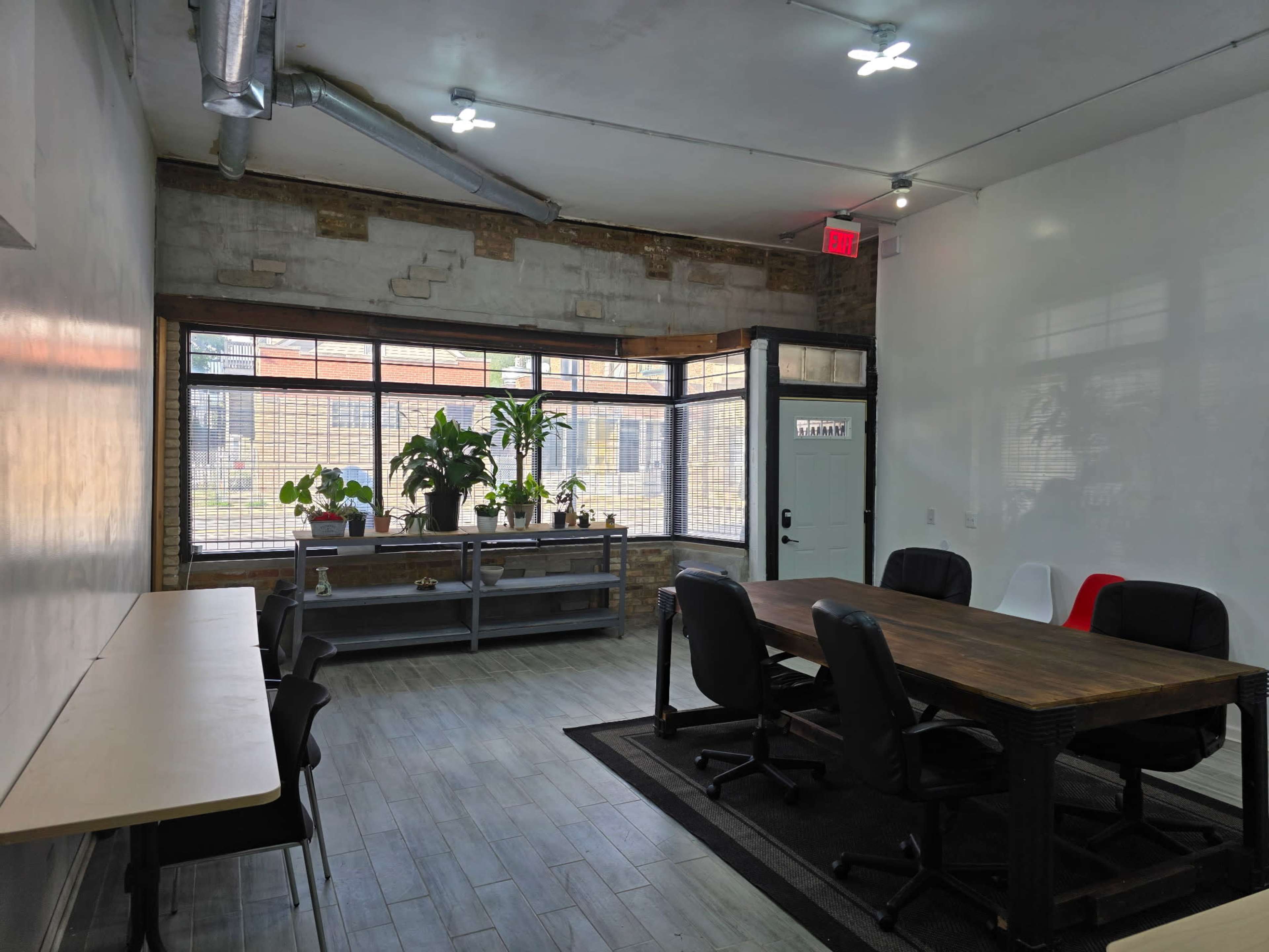 The image shows a spacious, well-lit office area with a large table surrounded by office chairs, a long desk along one wall, and large windows featuring potted plants.