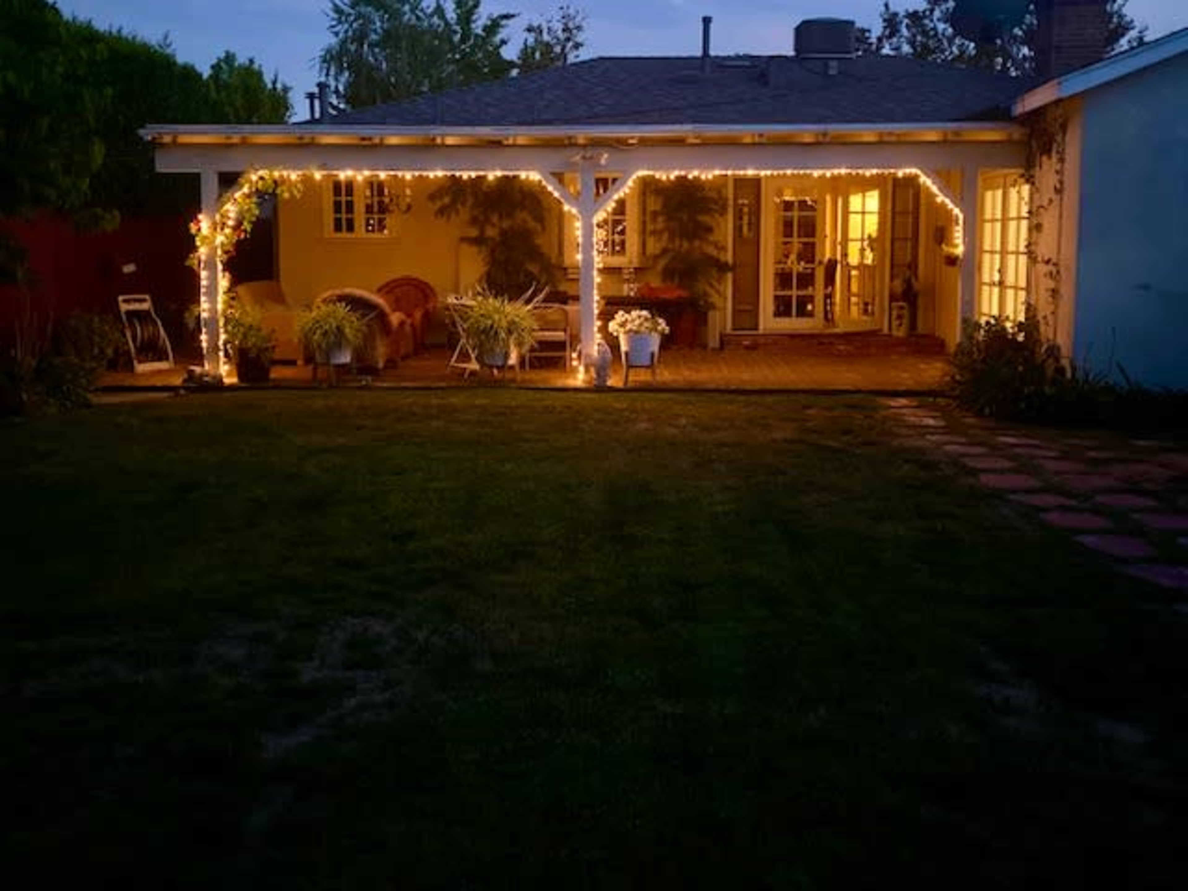 A cozy backyard is illuminated by string lights, showcasing a patio with potted plants and a view of an inviting house.