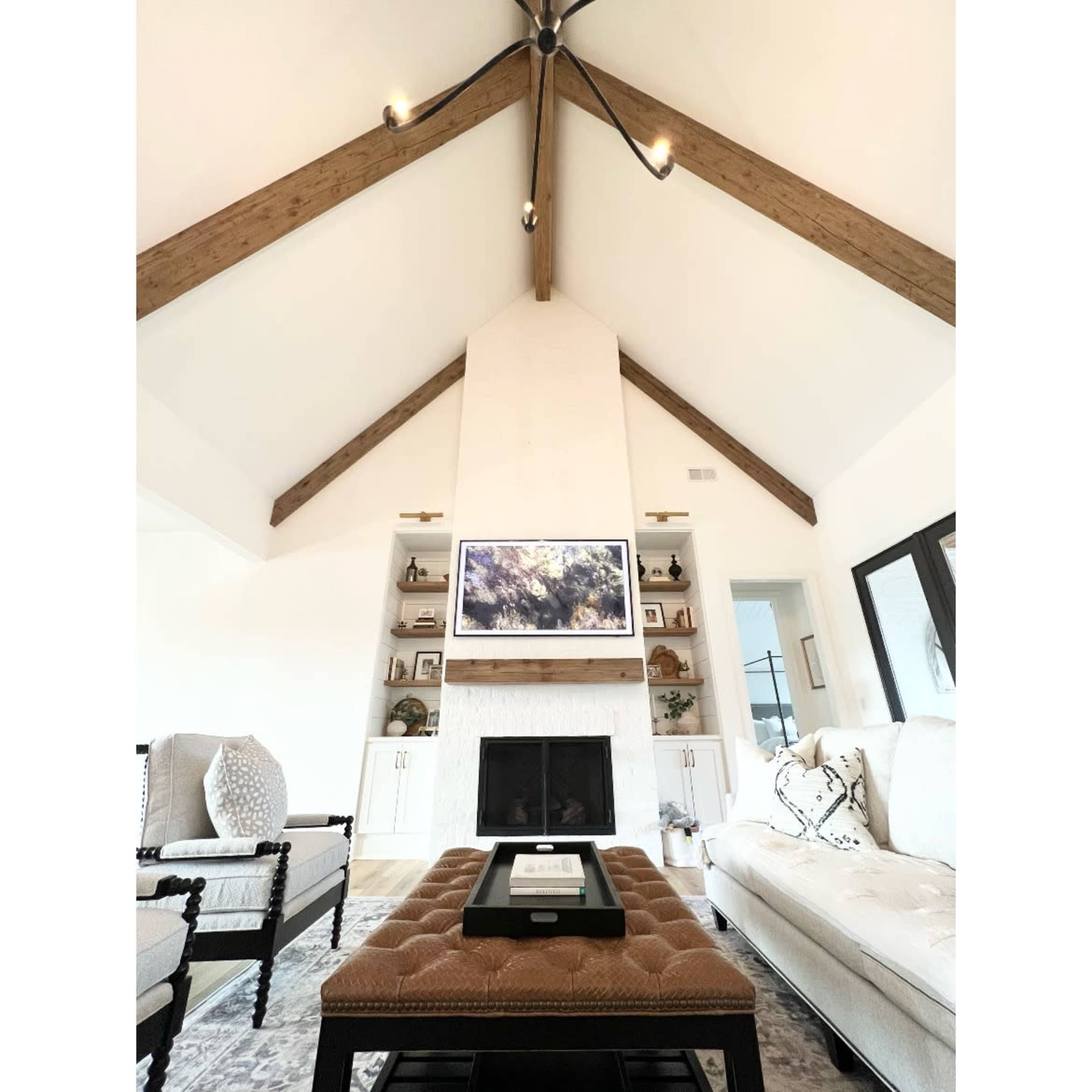 A living room with a high vaulted ceiling featuring wooden beams, a central fireplace with a television above it, and a coffee table in the foreground.