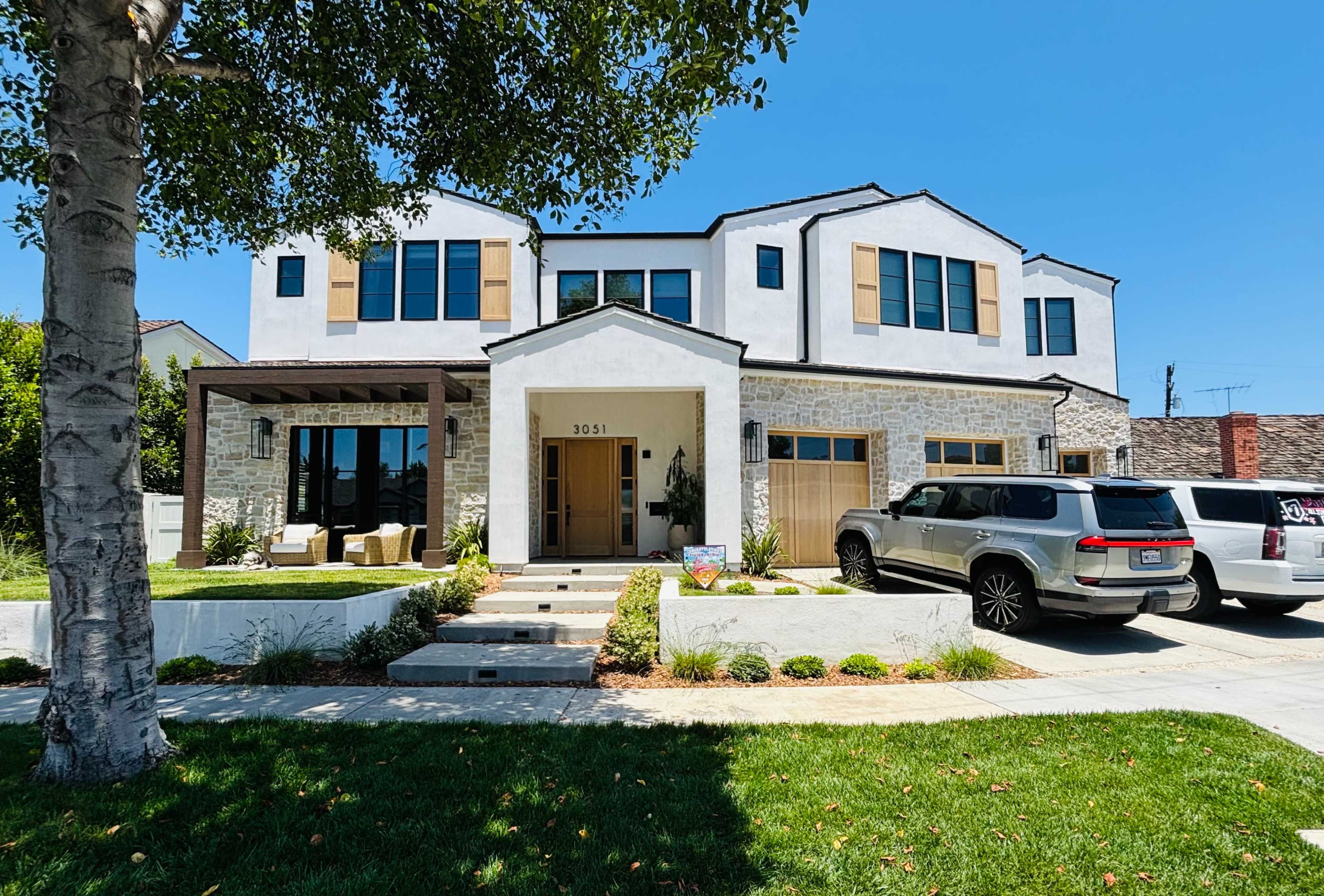 A modern two-story house with a stone facade, large windows, and a neatly landscaped front yard, featuring parked vehicles in the driveway.