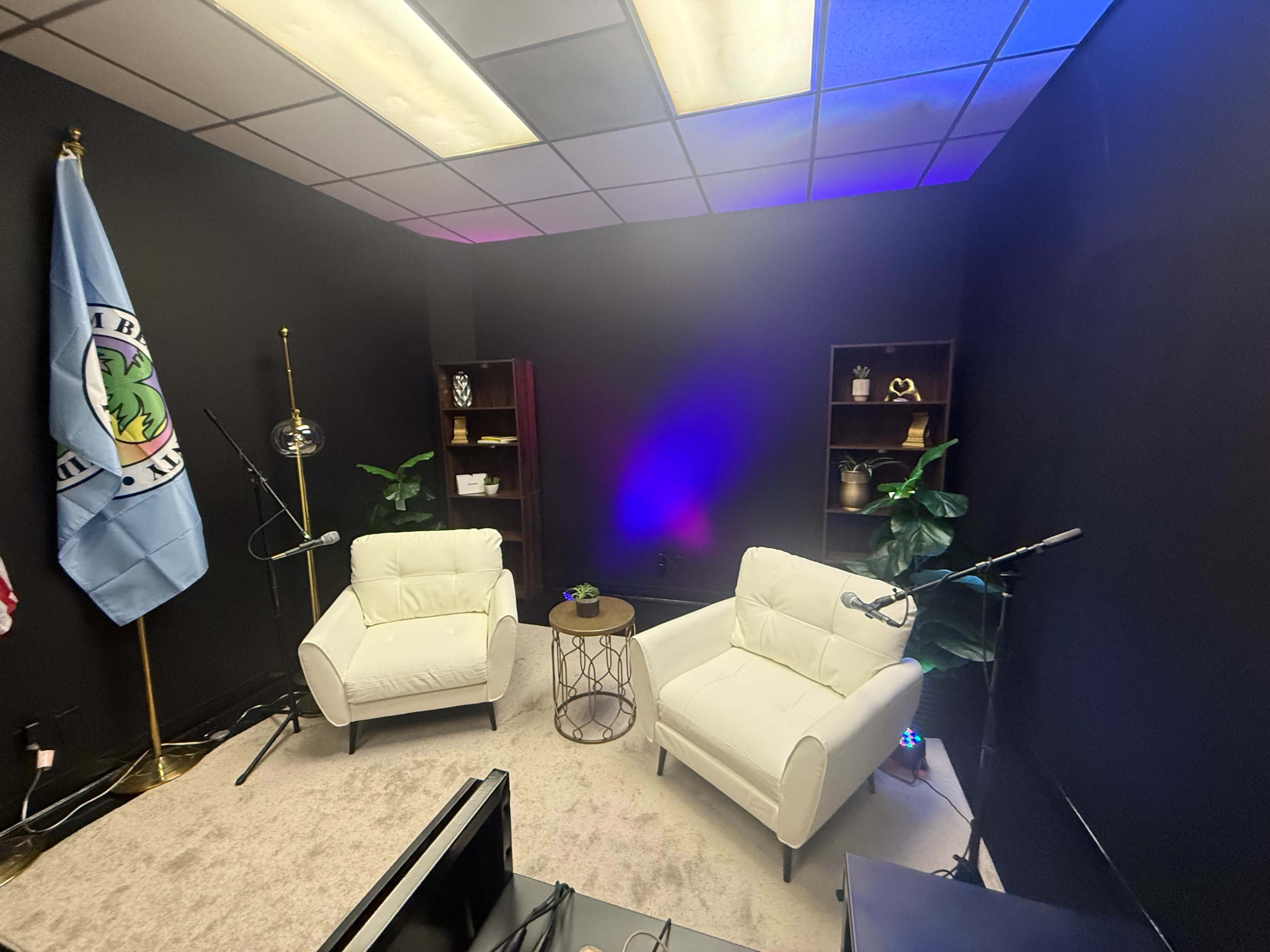 The image shows a small, neatly arranged studio space with two white armchairs, a side table, a microphone on a stand, a blue flag, and shelves with decorative items against a black wall.