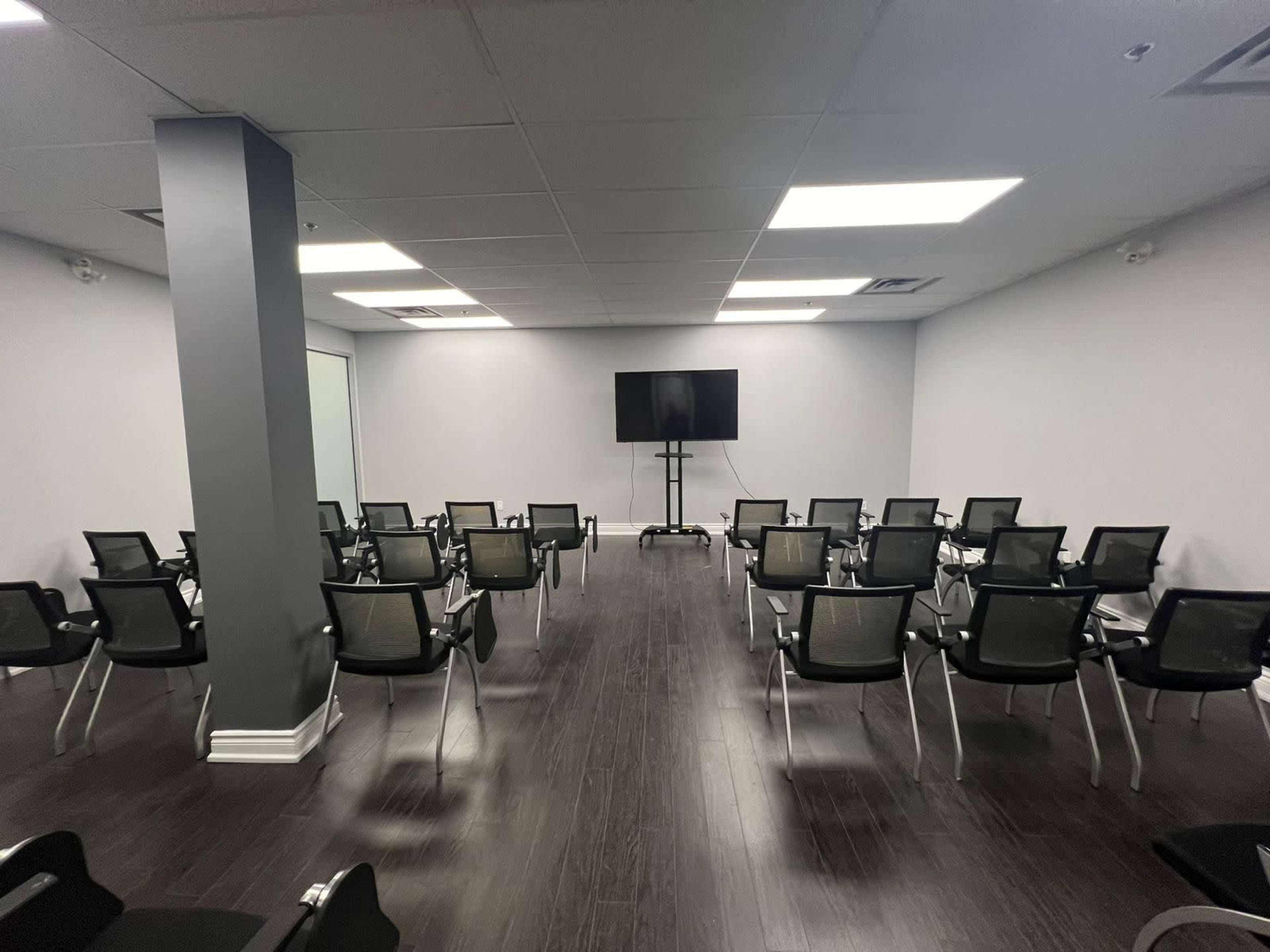 A conference room arranged with rows of black chairs facing a large wall-mounted screen.