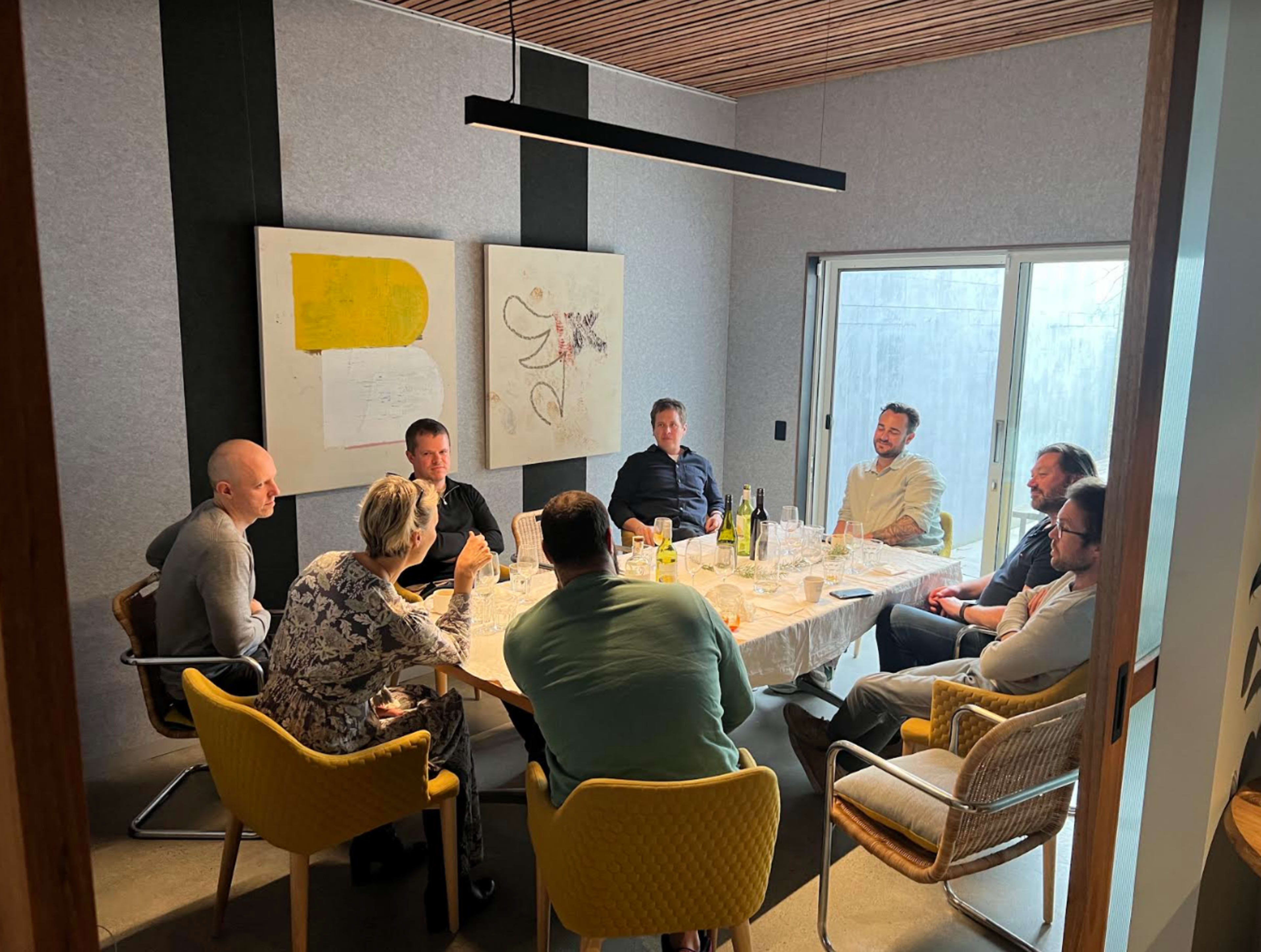 A group of eight people sits around a table in a modern room, engaged in conversation with various dishes and drinks in front of them.