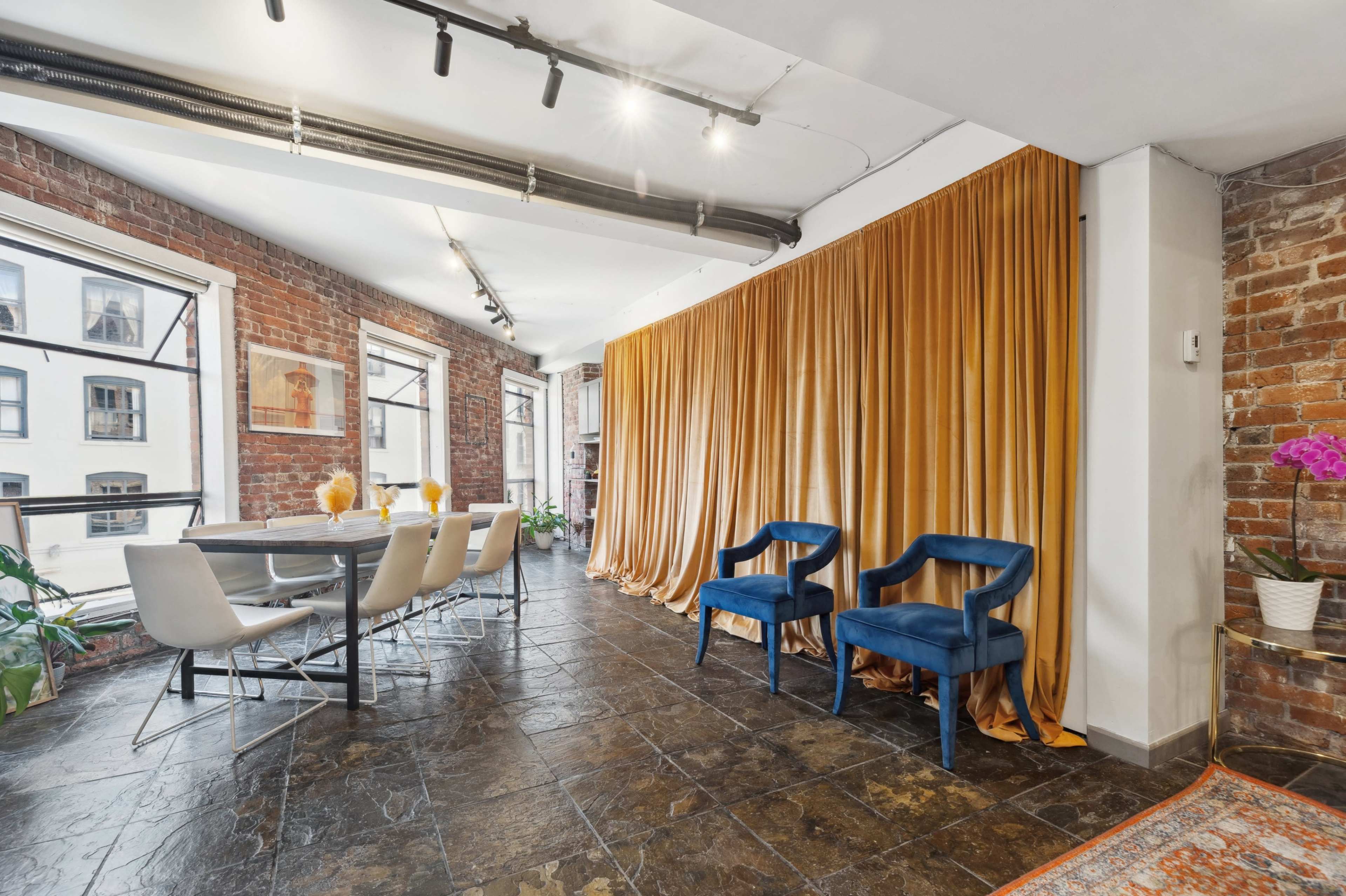 A modern dining area with a wooden table set for six, surrounded by exposed brick walls, and a curtain of golden fabric dividing the space.