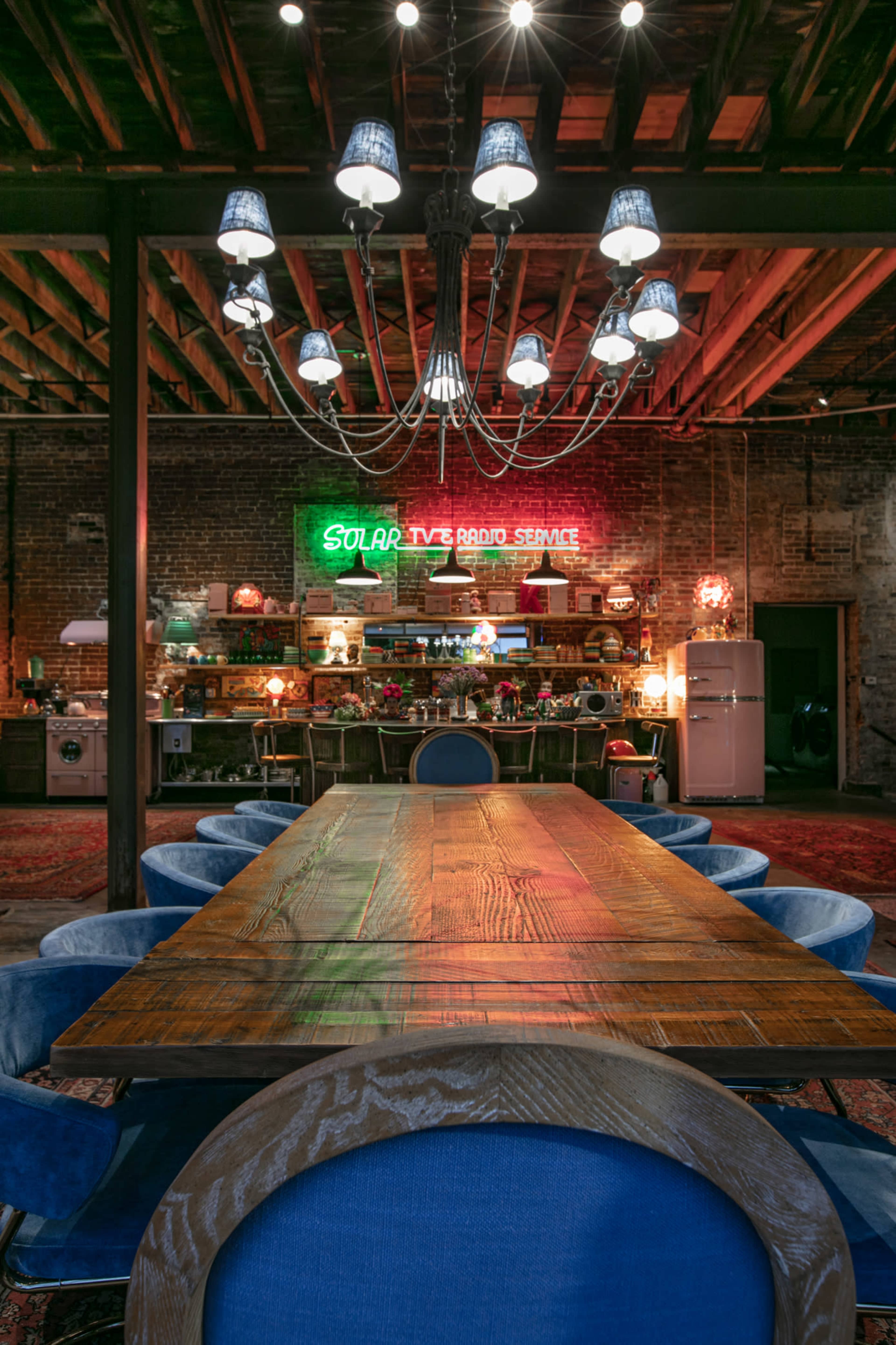 A large wooden dining table is set in a rustic room with exposed beams and a neon sign that reads "Solar TV Radio Service" in the background.