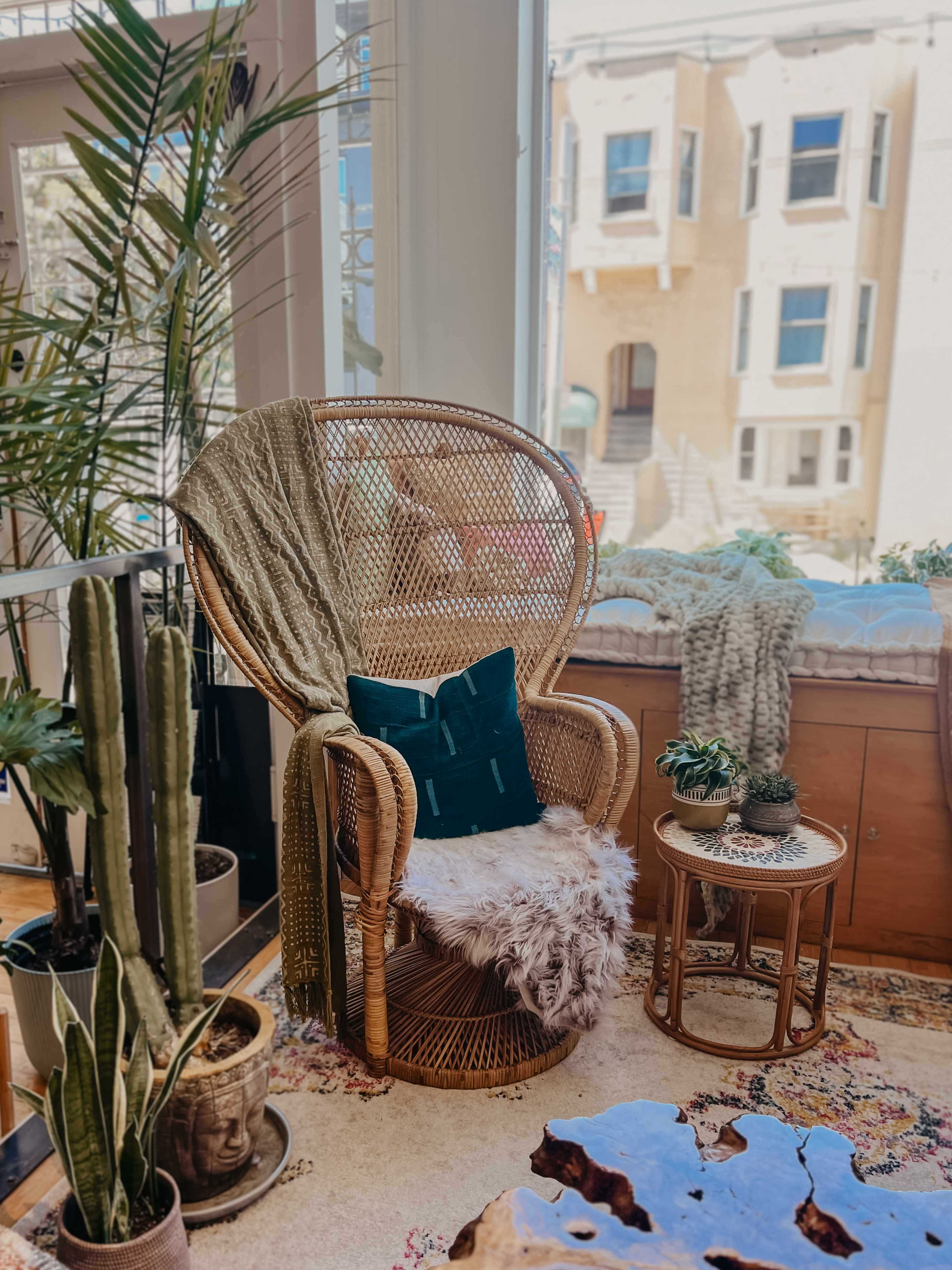 A rattan chair with a green pillow and a faux fur throw is positioned near a window, surrounded by potted plants and a small table with a plant.