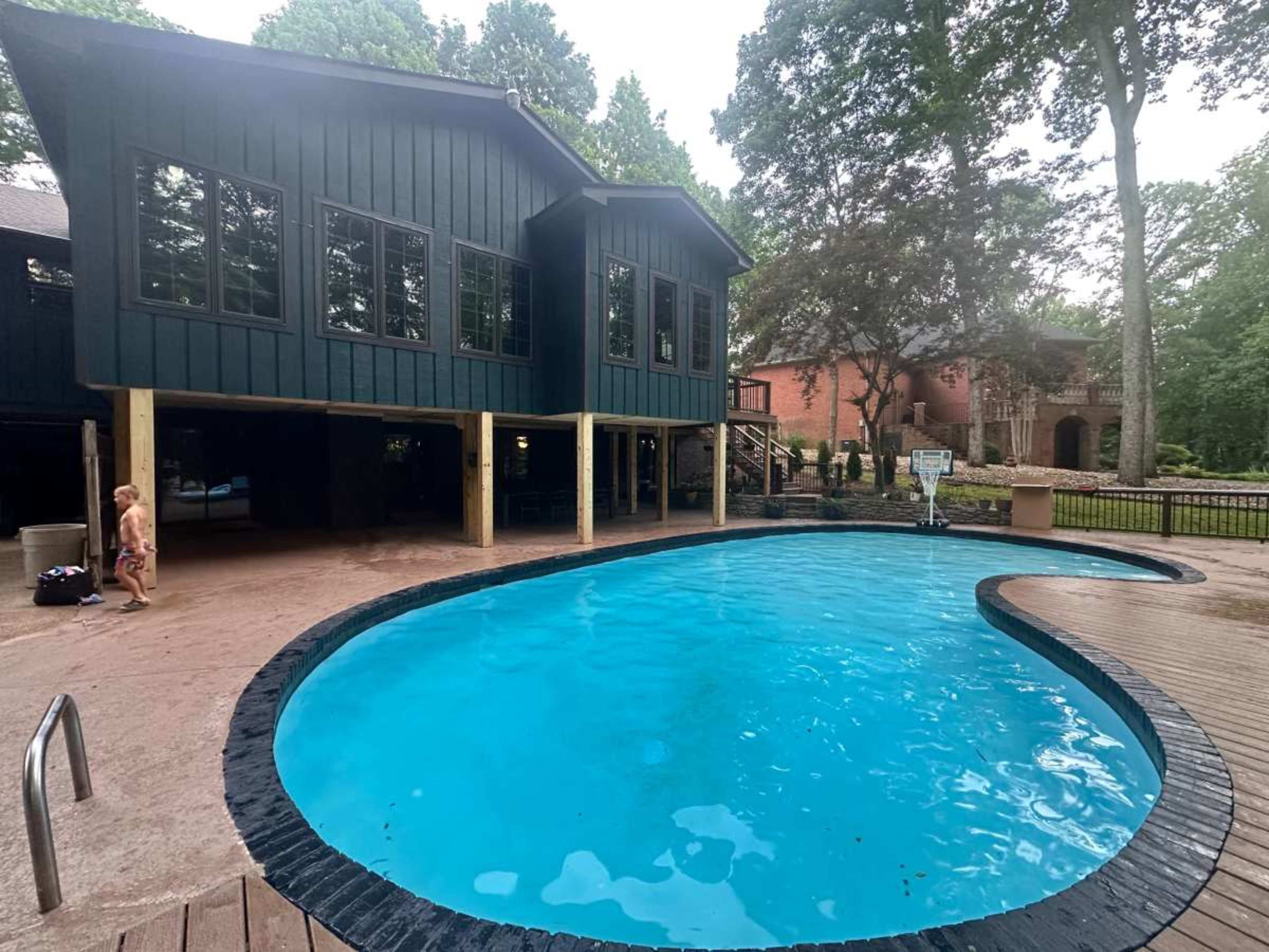 The image shows a blue swimming pool surrounded by a wooden deck, with a two-story dark green house and a large brick building in the background.