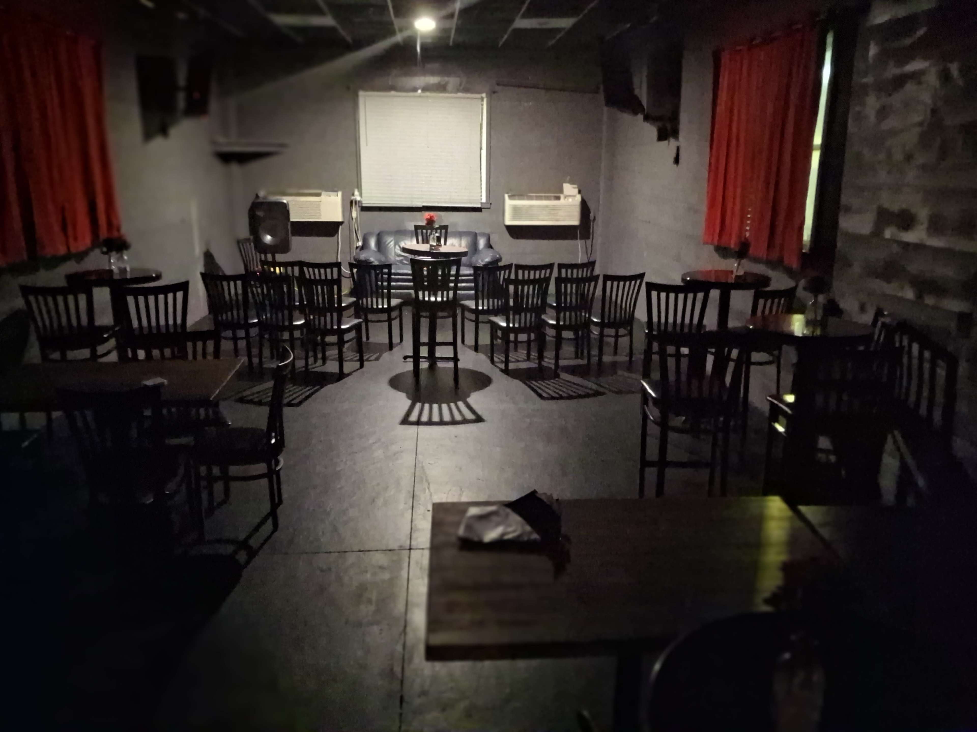 The image shows a dimly lit lounge area with rows of chairs and tables, flanked by red curtains and a blank screen at the front.