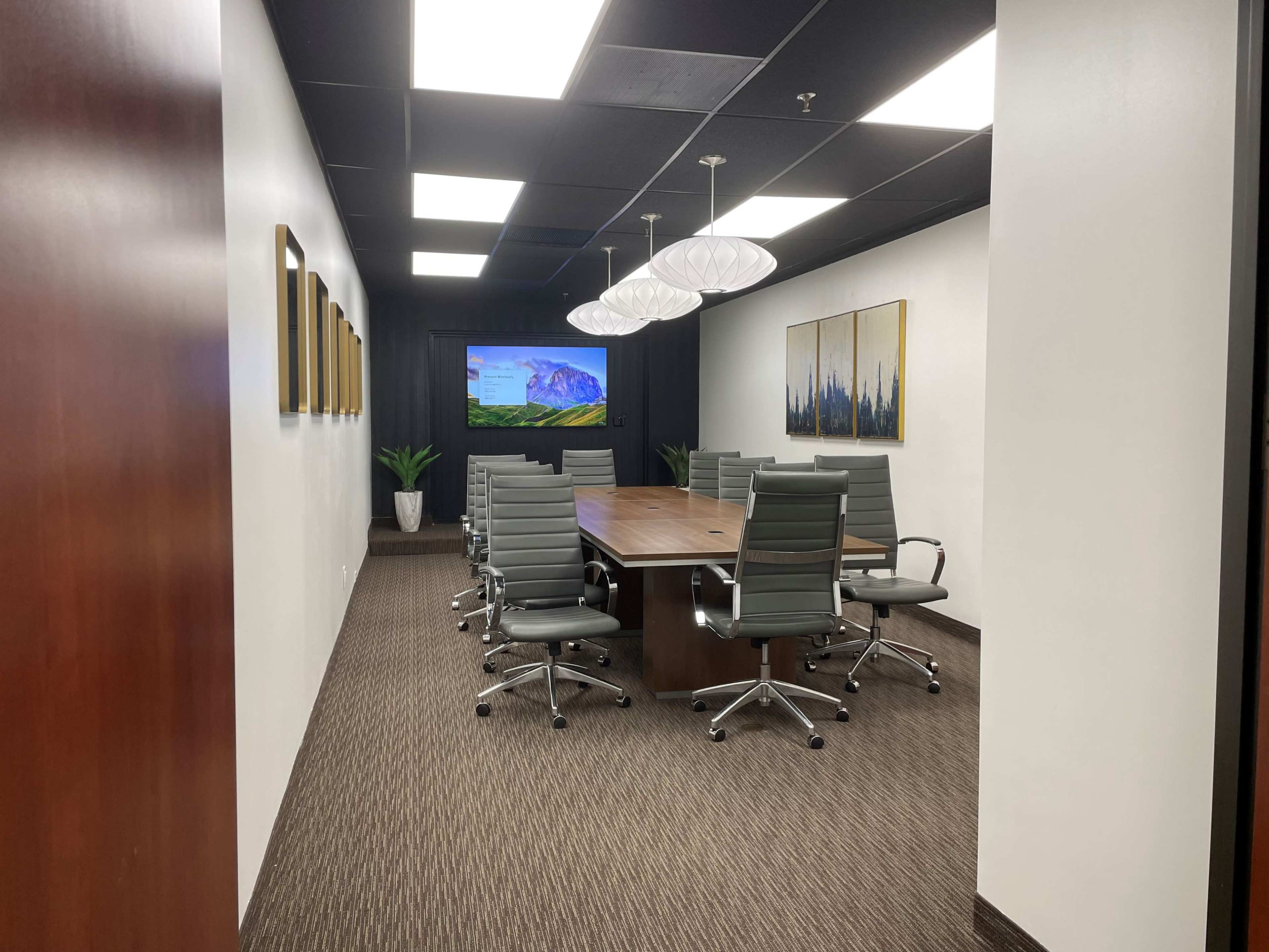 The image shows a modern conference room with a large wooden table, six gray leather chairs, a mounted screen displaying a landscape, and artwork on the walls.