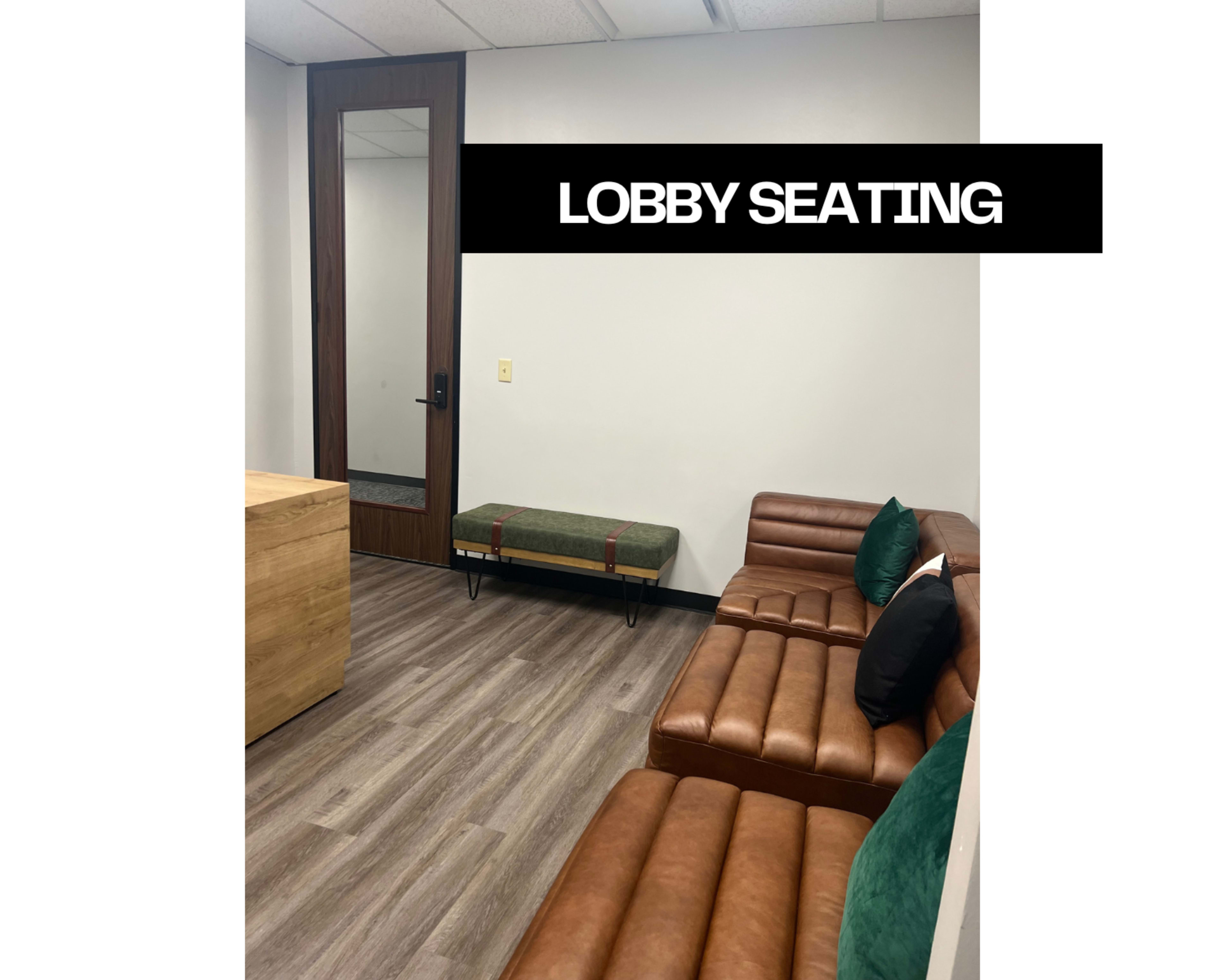 The image shows a lobby seating area featuring a brown leather couch, a green bench, and a wooden table, with a mirror and door visible in the background.