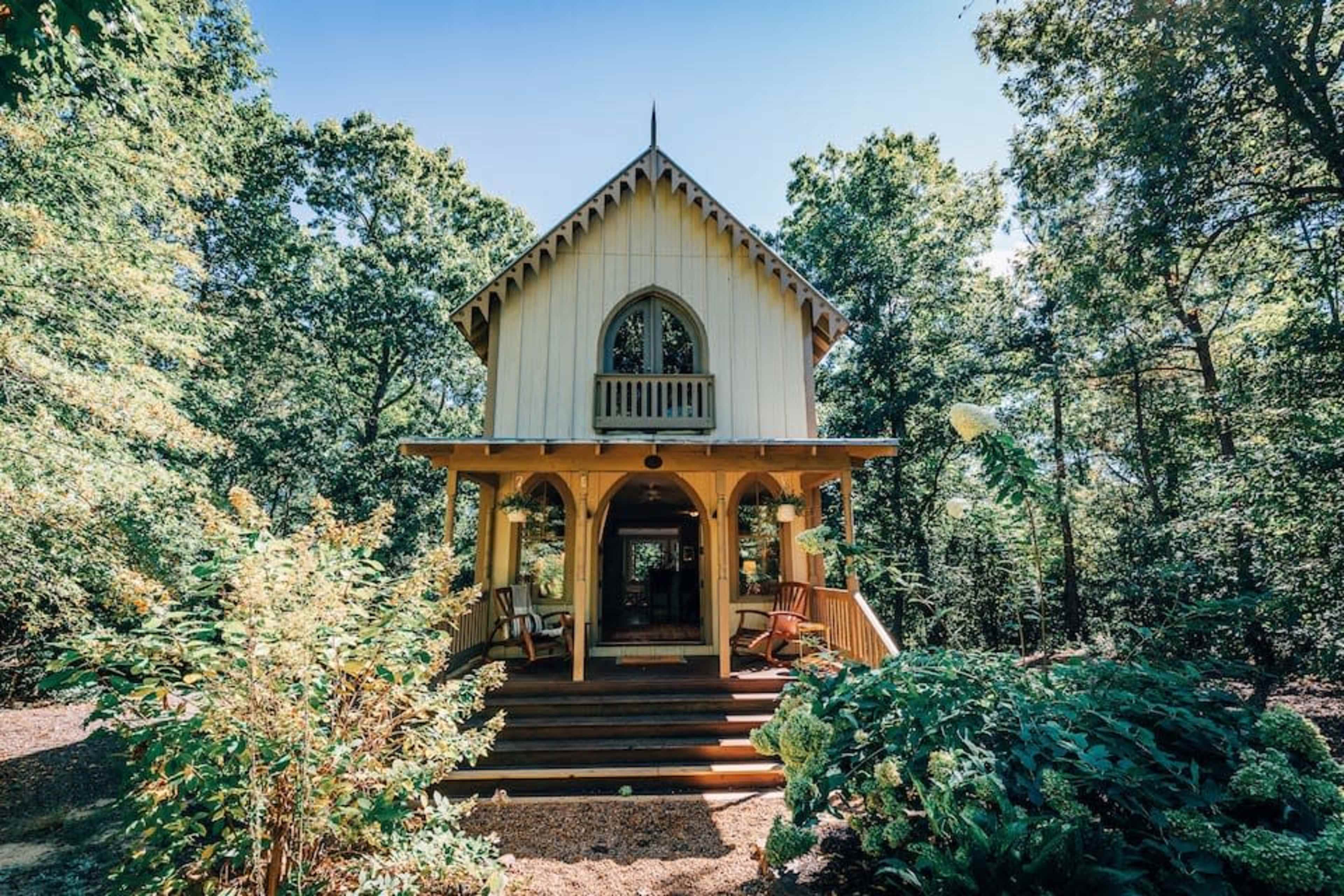 A two-story cottage with a peaked roof and balcony is surrounded by trees and greenery.