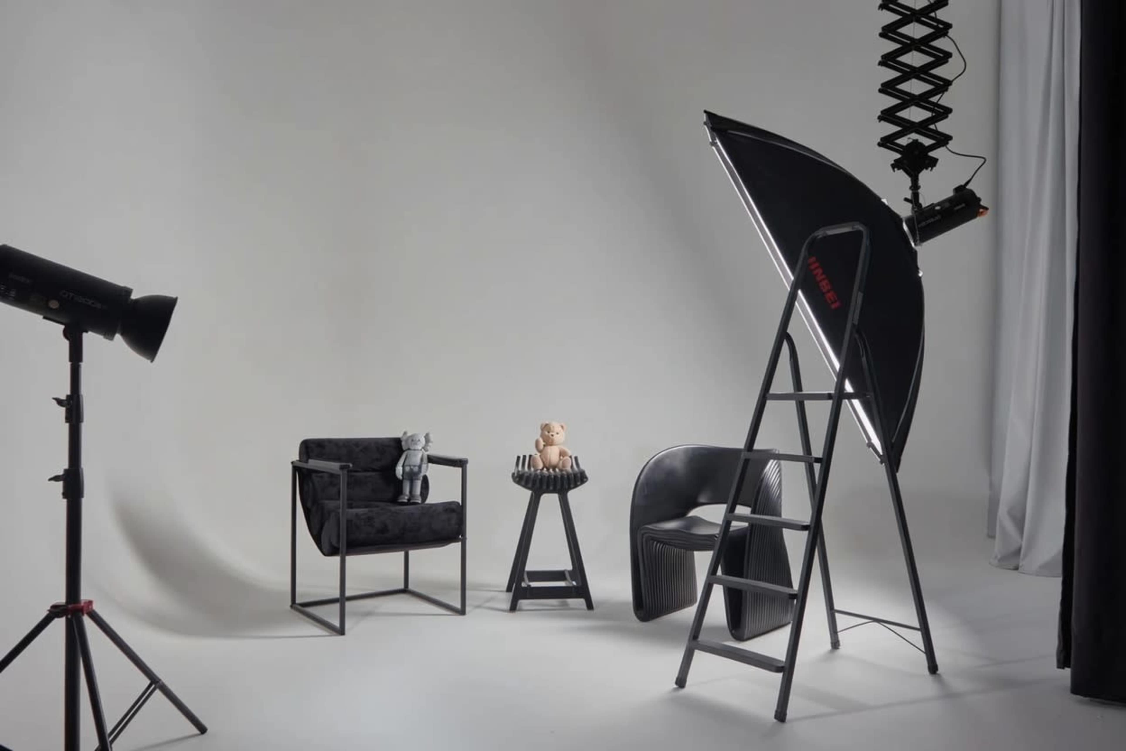 The scene features a minimalist photography studio setting with two chairs, a small table with decorative items, and studio lighting equipment.
