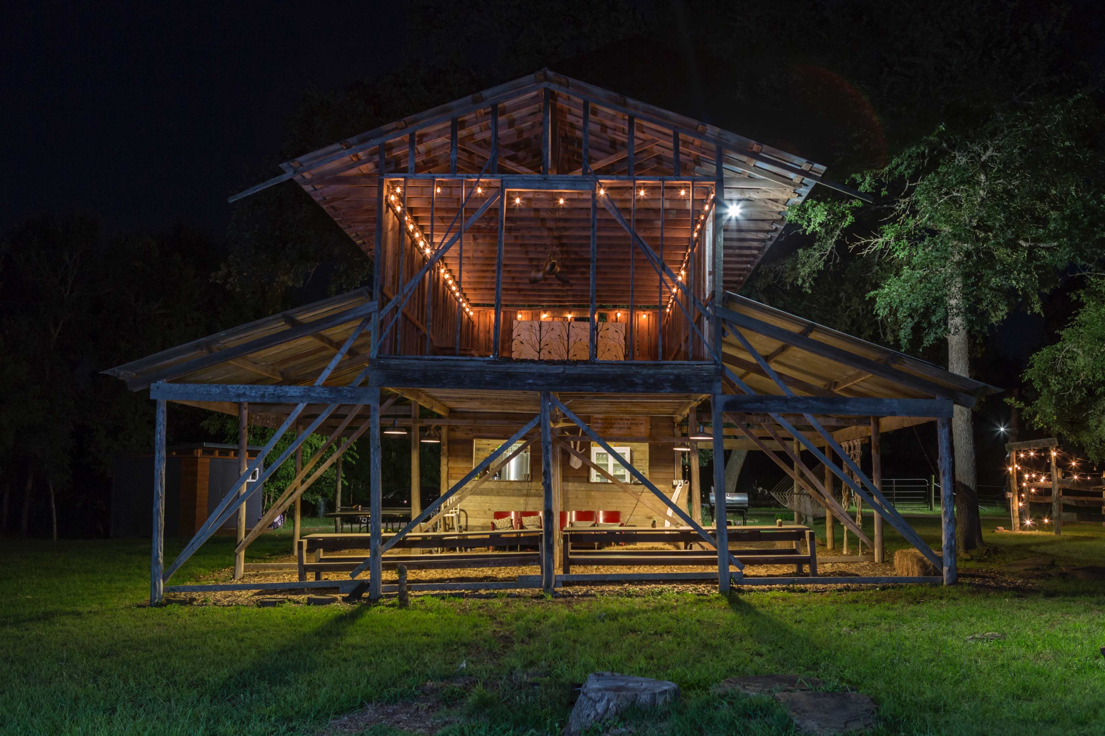 A wooden structure with an open lower level and a lofted upper area, illuminated by string lights at night, stands in a grassy environment.