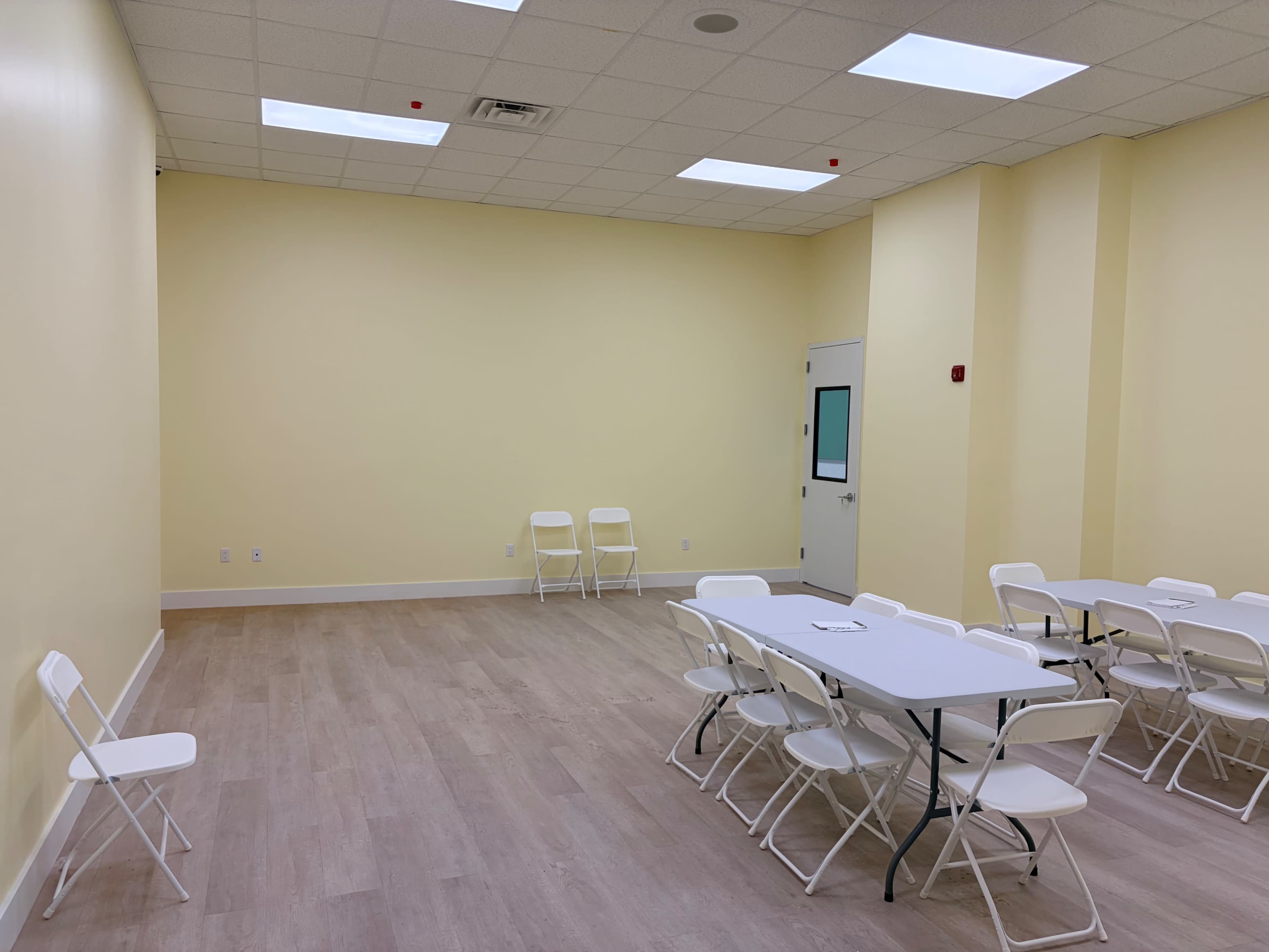 The image depicts a sparsely furnished room featuring a light yellow wall, several white folding chairs, and a rectangular table arranged in the center.