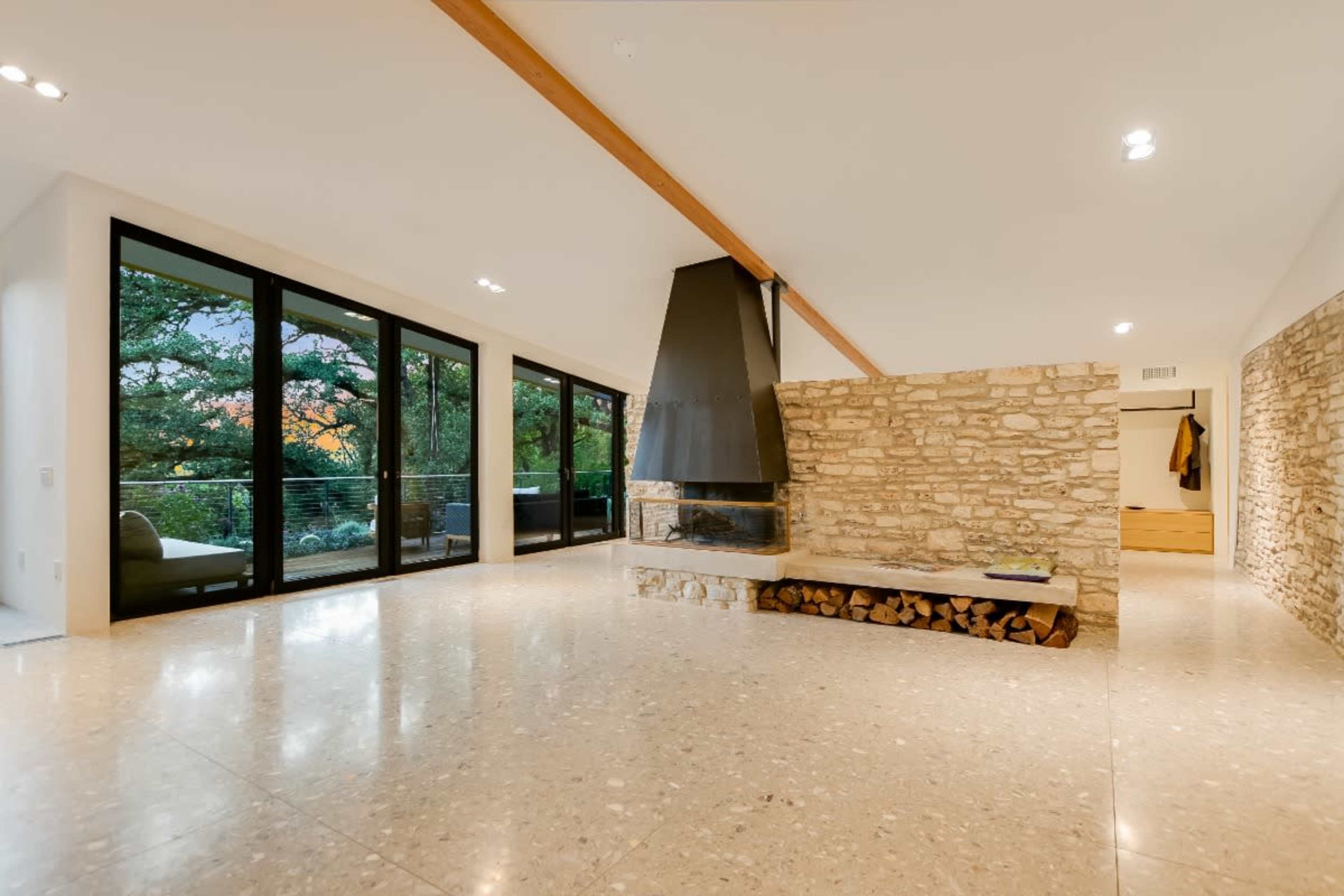 The image shows a modern living space with a stone fireplace, large windows overlooking greenery, and polished flooring.