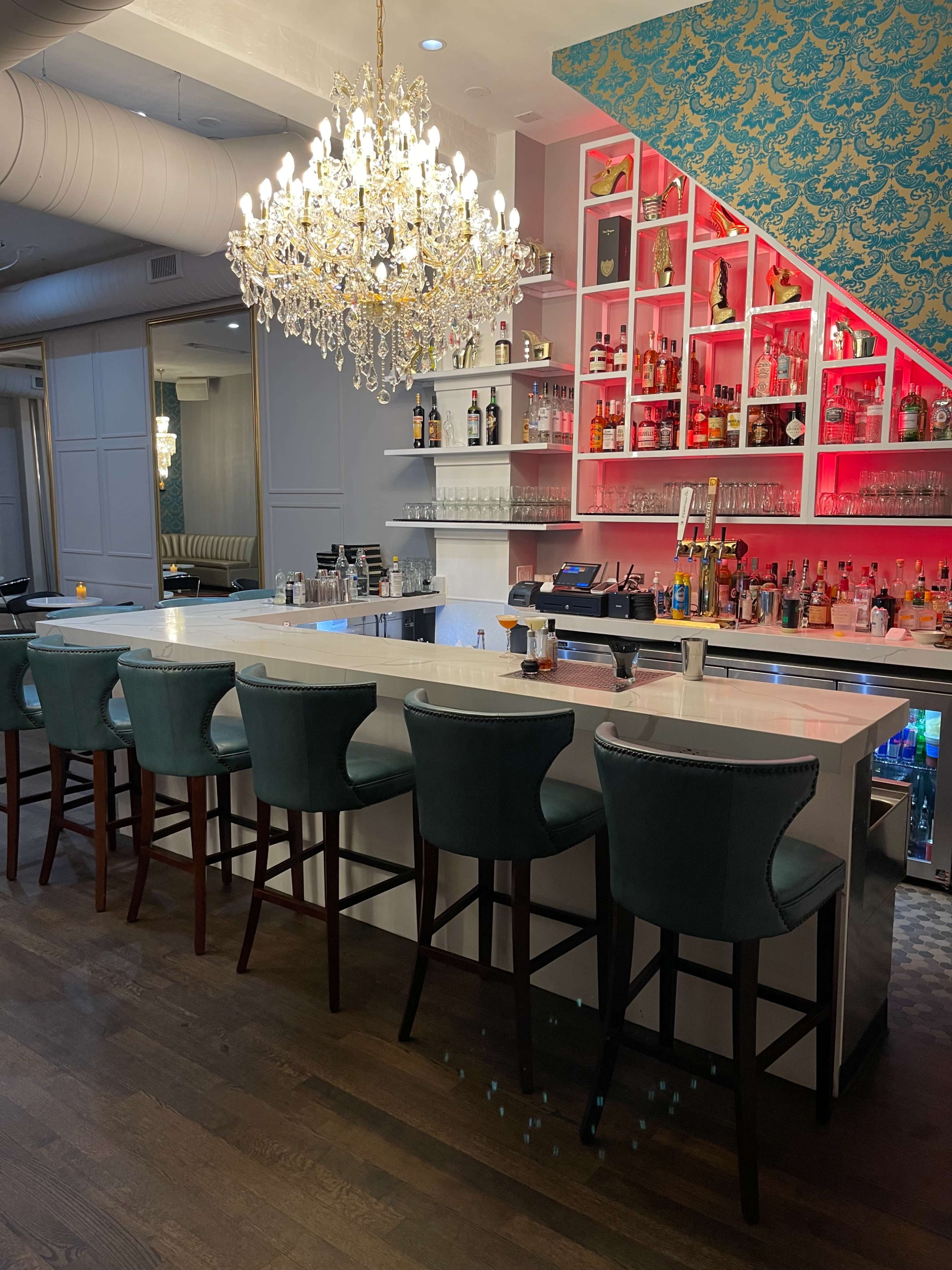 A modern bar with a white countertop, turquoise upholstered stools, and a sparkling chandelier above, against a backdrop of shelves filled with liquor bottles.