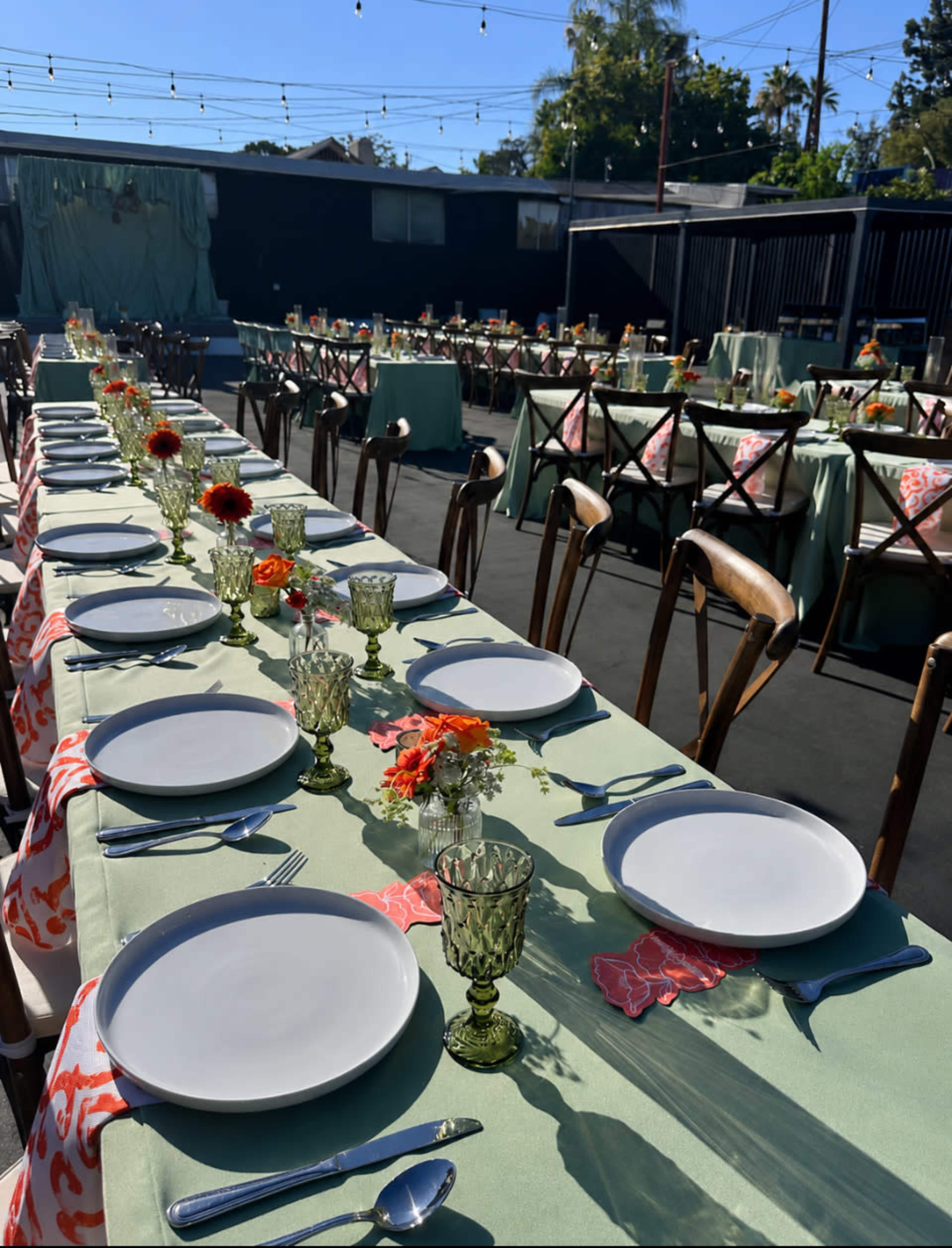 A long outdoor dining setup features multiple tables adorned with light green tablecloths, white plates, decorative glassware, and floral centerpieces.