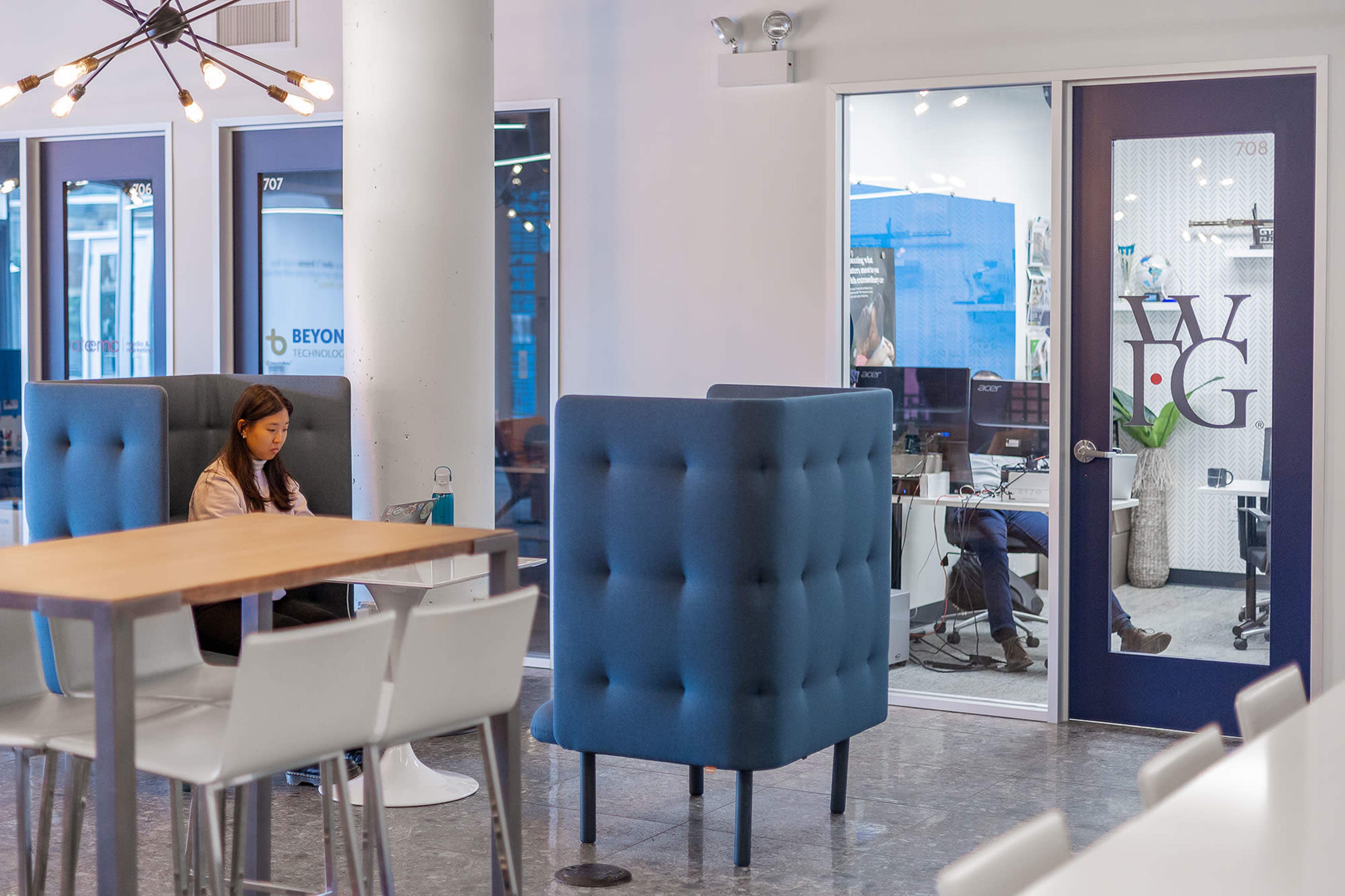 A woman sits at a table using a laptop in a modern office space, while two people are visible inside an office behind a glass door.