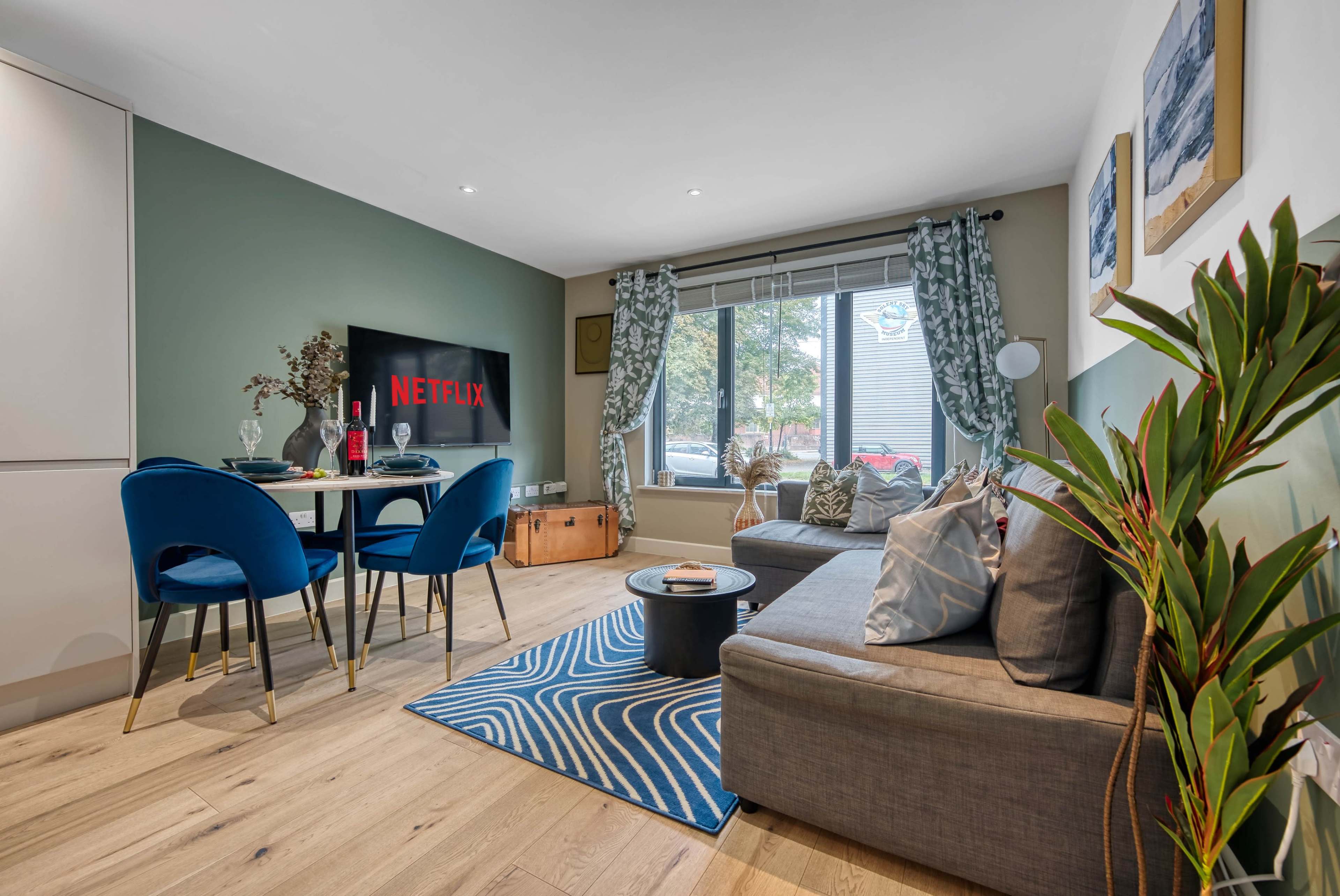 The living room features a sofa, a round table with blue chairs, and a television displaying the Netflix logo, with large windows allowing natural light.