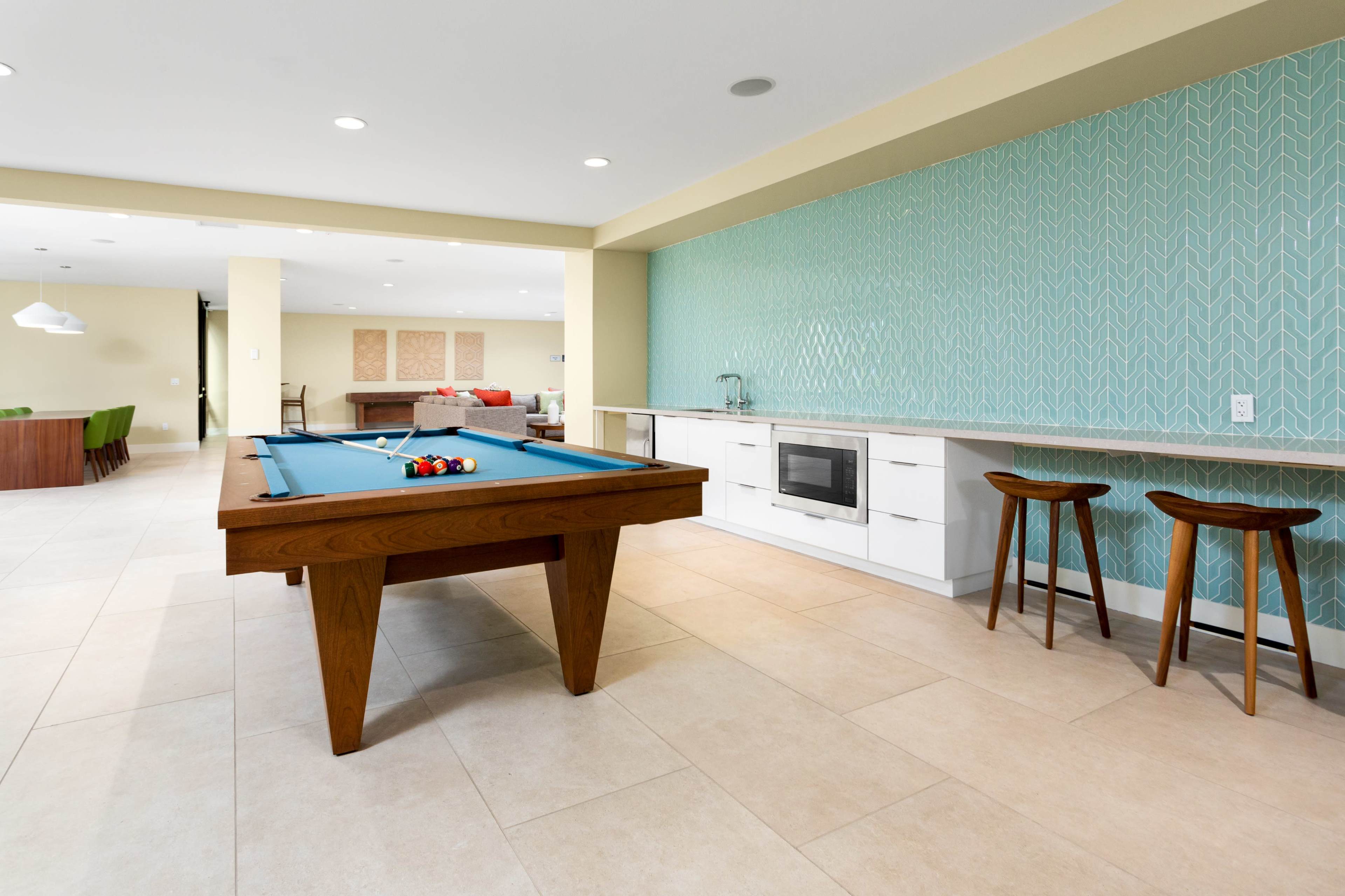The image shows a modern recreational space with a blue pool table in the foreground, a kitchen area in the background, and stylish seating arrangements.