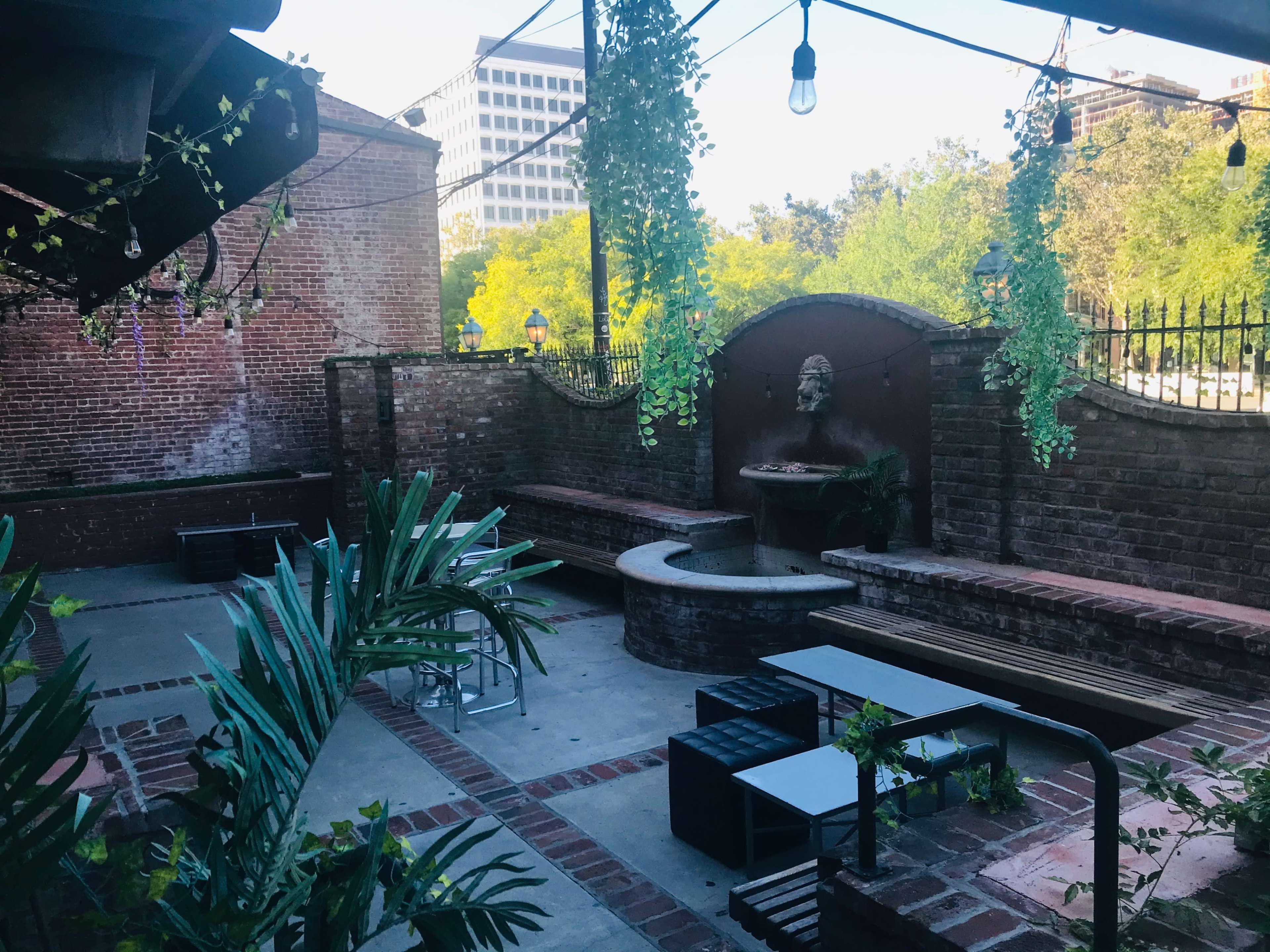 The image shows a brick-walled outdoor courtyard with a fountain, seating areas, and hanging plants.