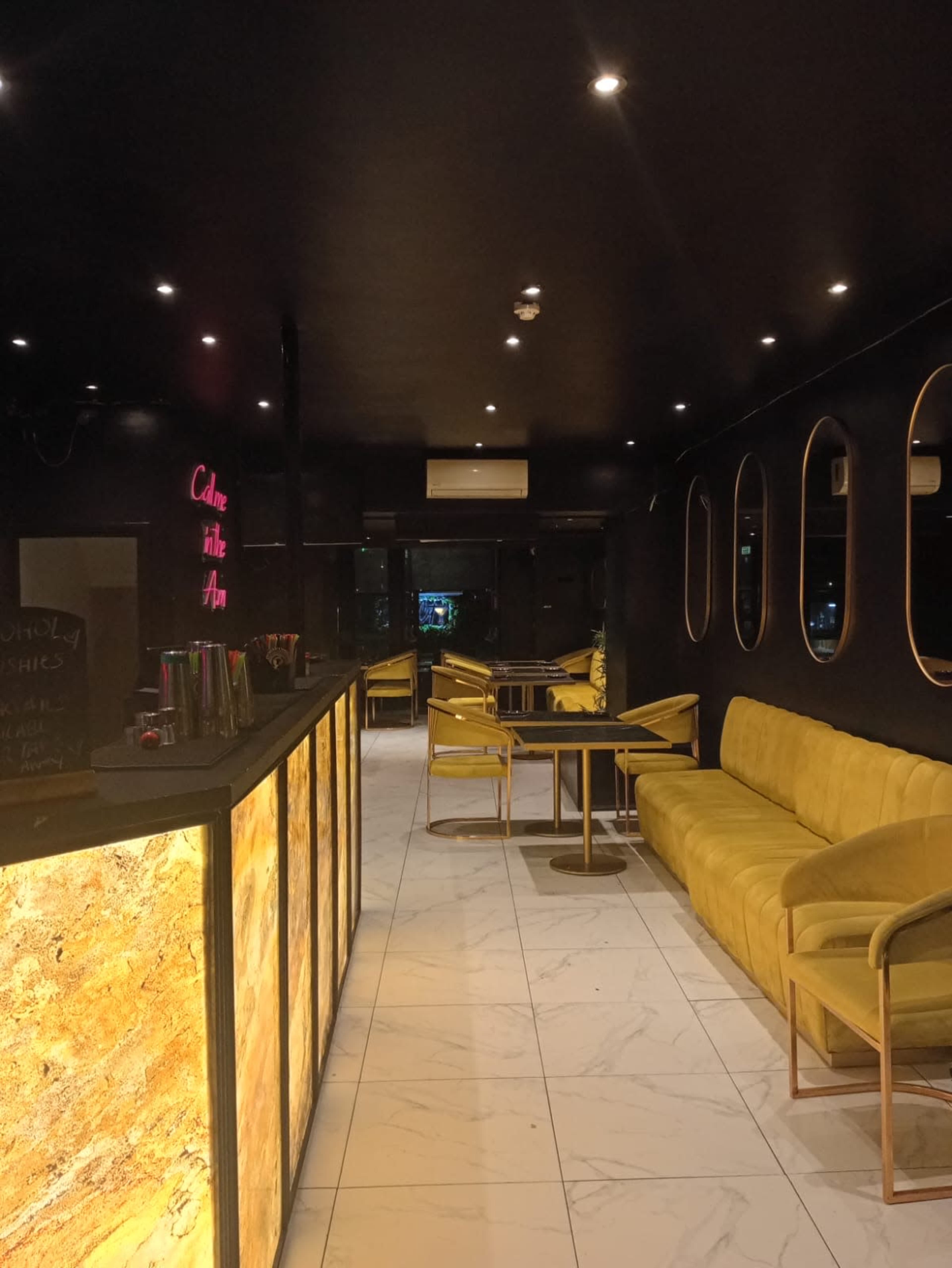 The image shows a modern, dimly lit café with yellow seating and a bar counter made of textured material, featuring circular mirrors on the walls.