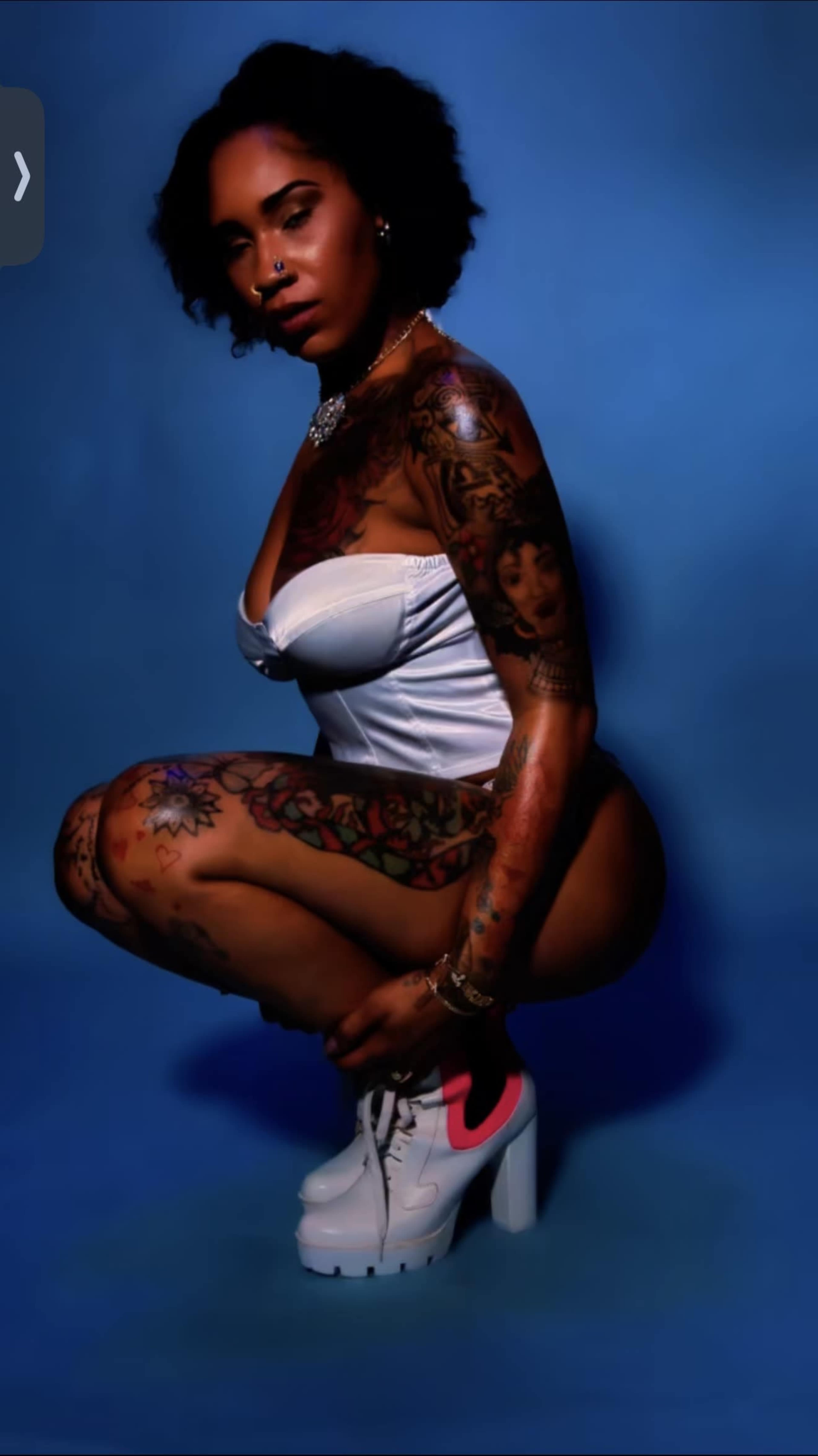 A woman with extensive tattoos is squatting against a blue background while wearing a white top and a single pink-accented shoe.