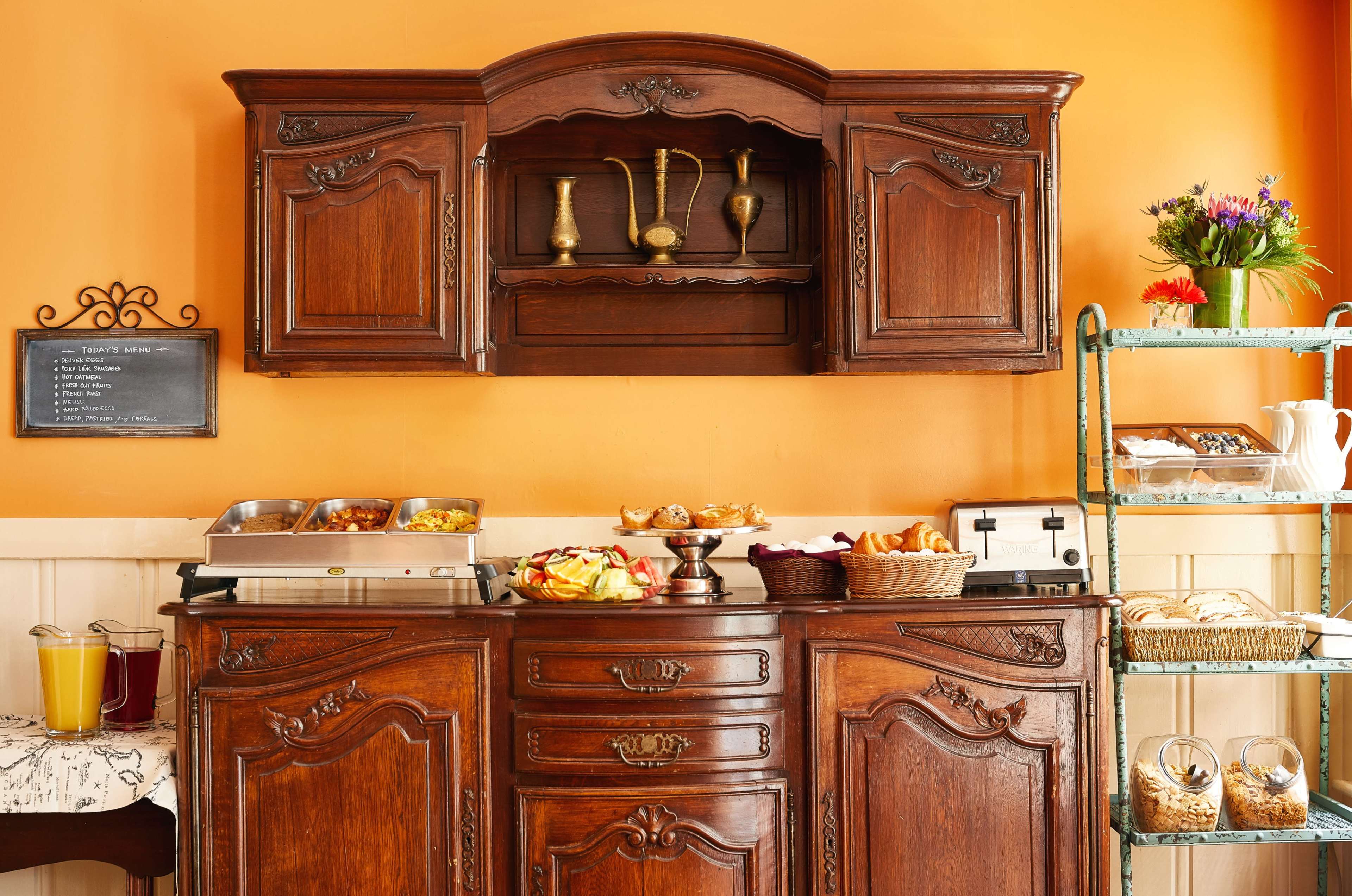 A wooden buffet table with a display of food items is set against an orange wall, with a shelf holding decorative pieces above it.