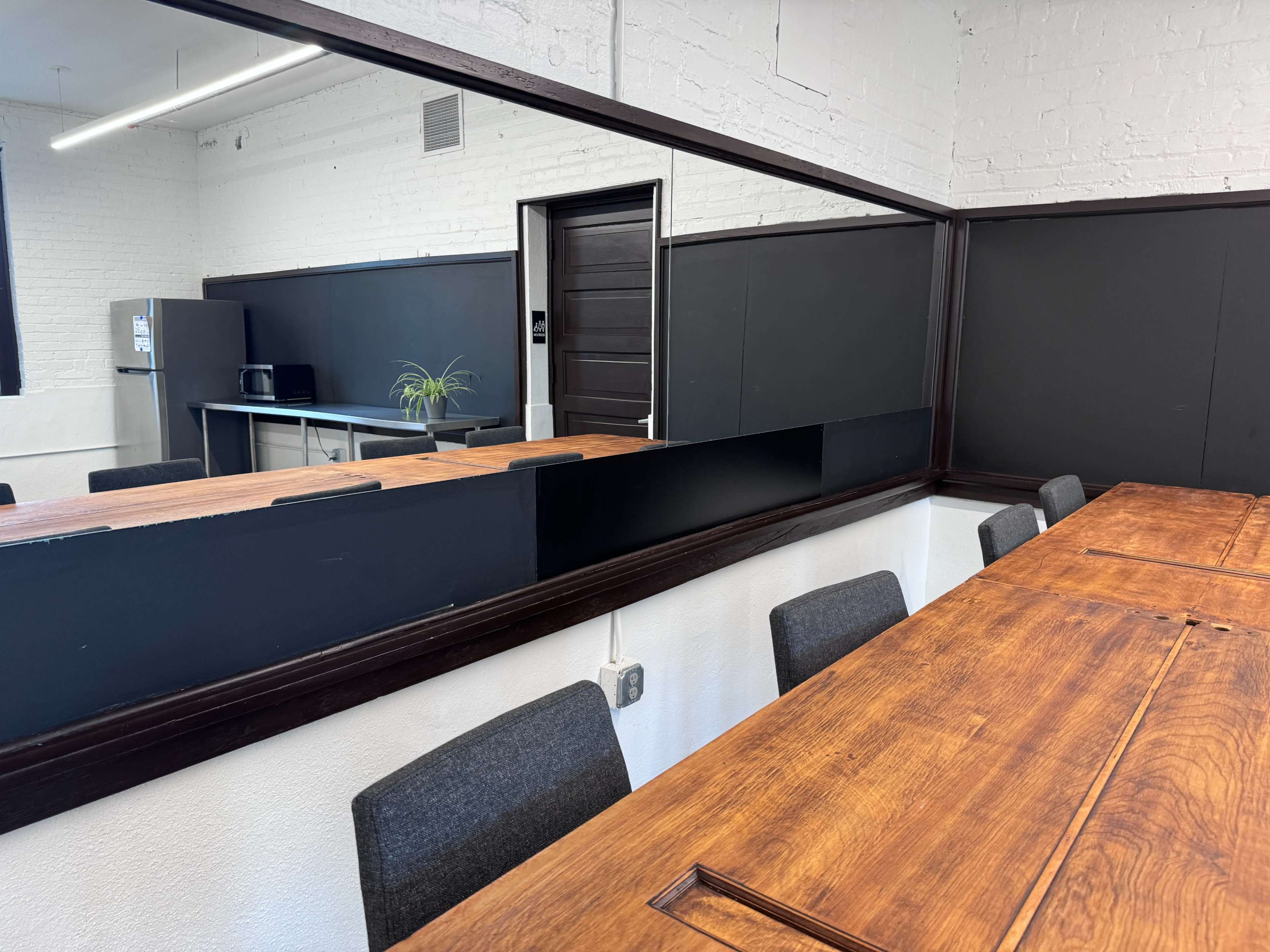 A large wooden table with gray chairs is positioned in a meeting room that features a mirrored wall, a kitchenette in the background, and black paneling.