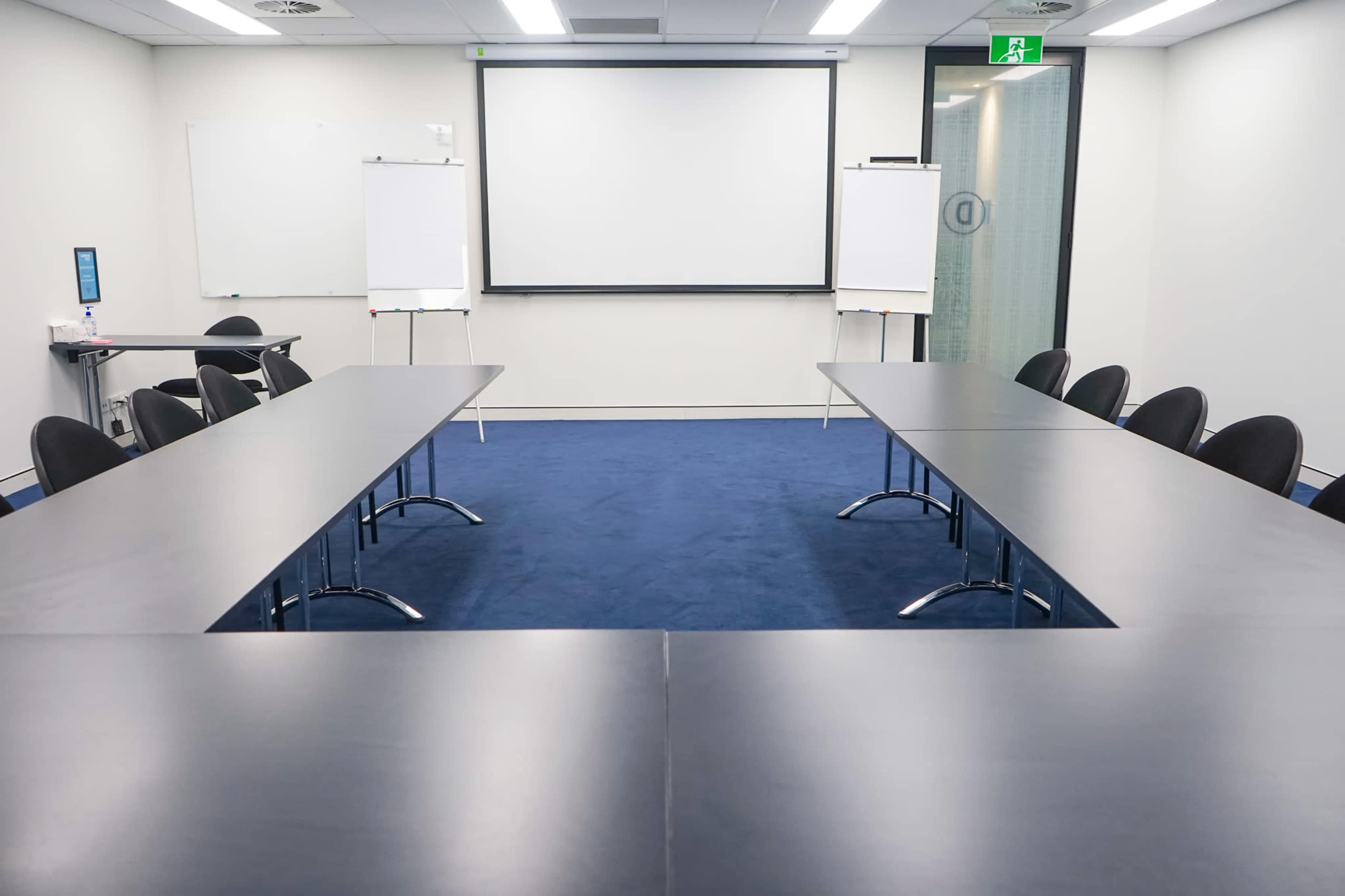 A conference room is set up with two rectangular tables facing a presentation screen, flanked by empty chairs and whiteboards.