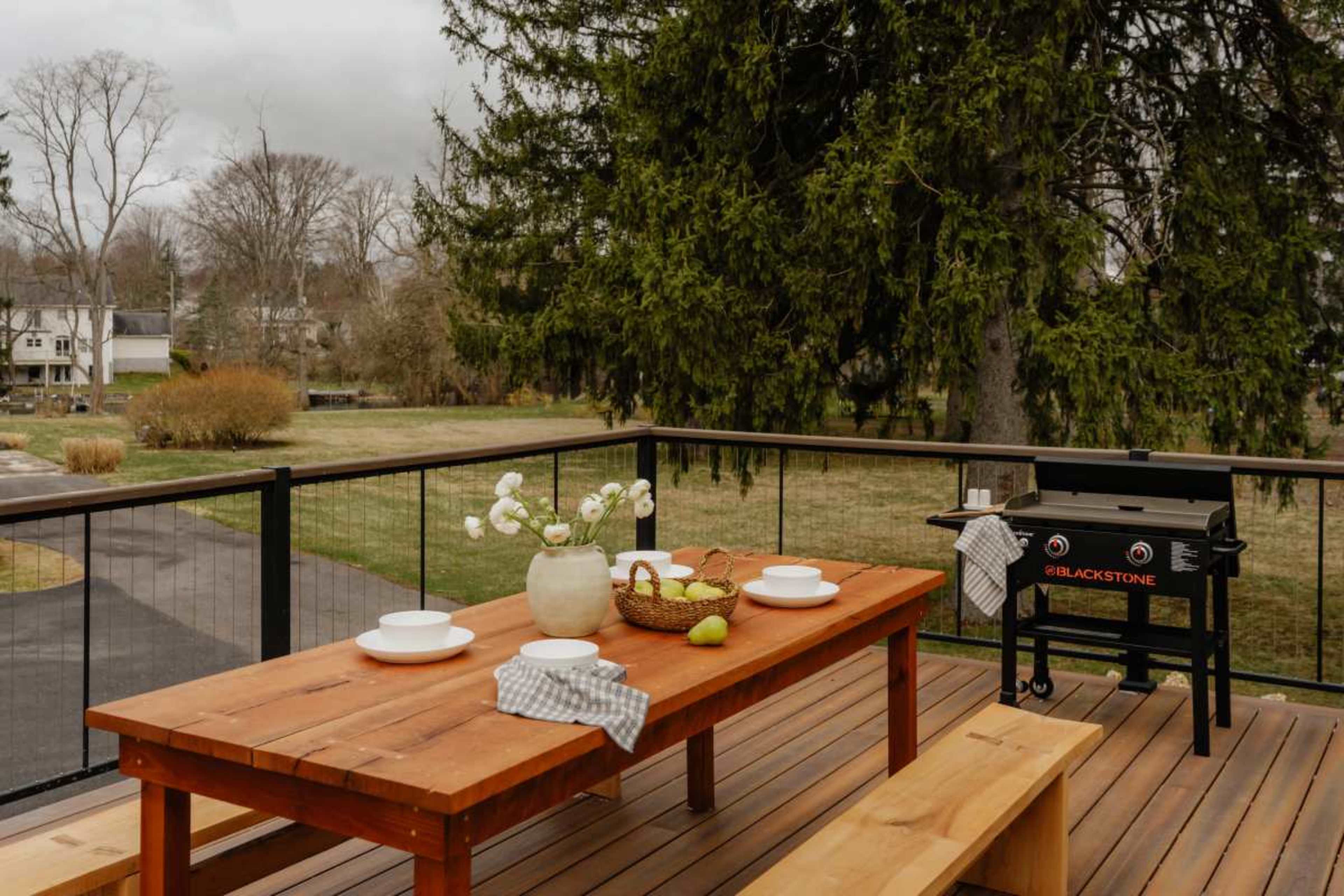 A wooden dining table is set with plates and a vase of flowers on a deck beside a Blackstone grill, overlooking a grassy yard and trees.