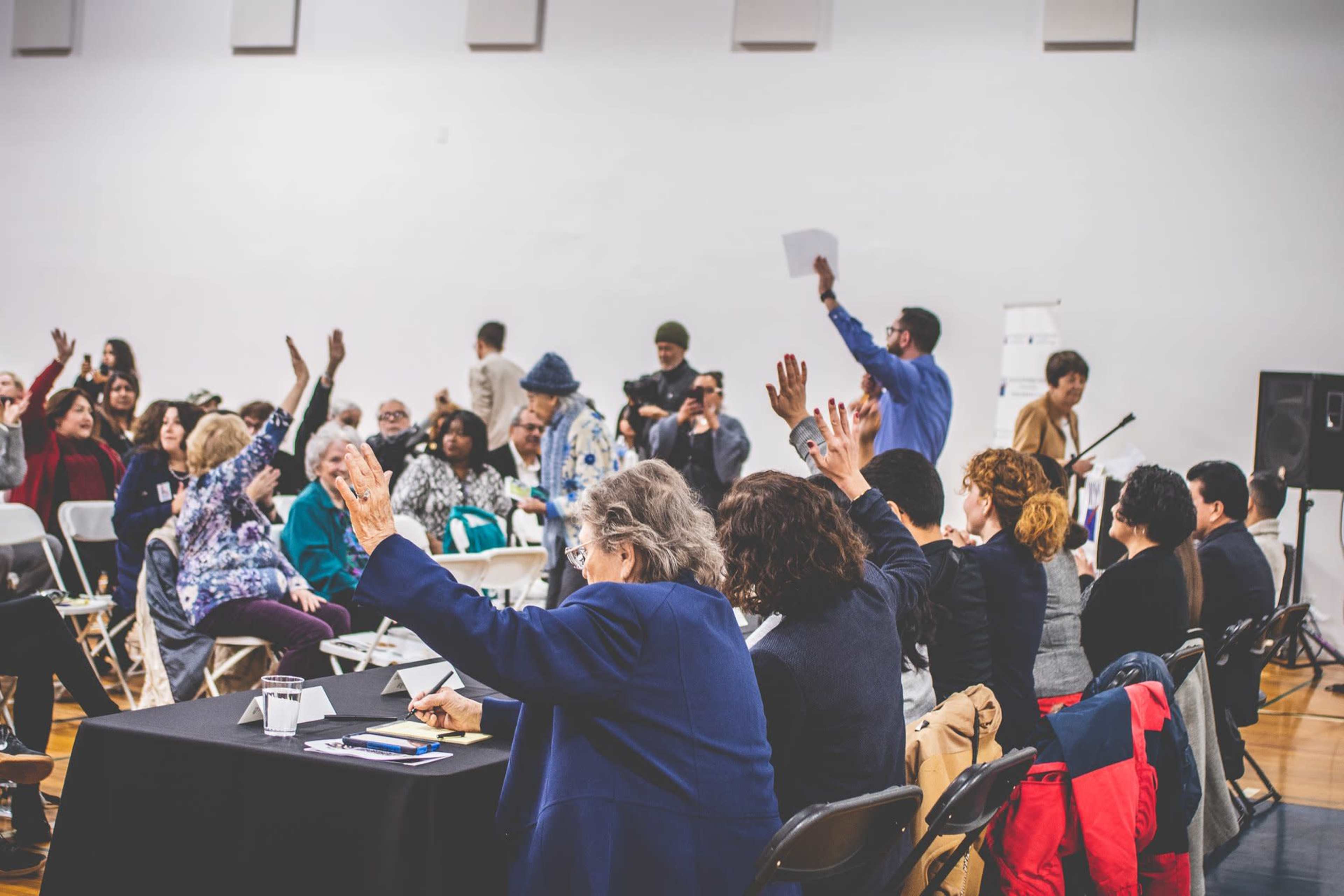 A group of people raises their hands in a large meeting space, while someone stands at the front holding a sheet of paper.