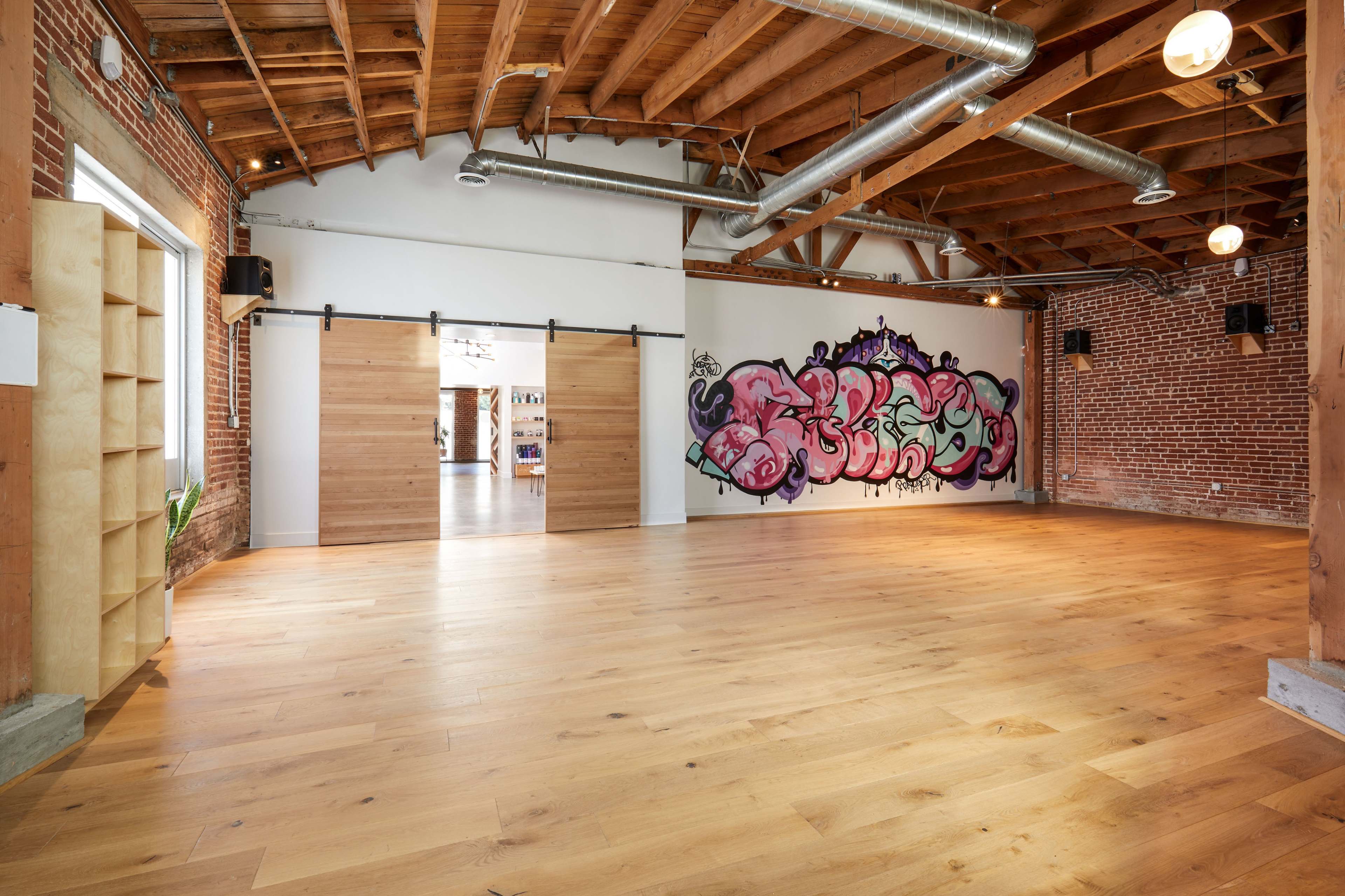 A spacious interior room with wooden floors, exposed brick walls, and a large mural on one wall, complemented by sliding wooden doors.