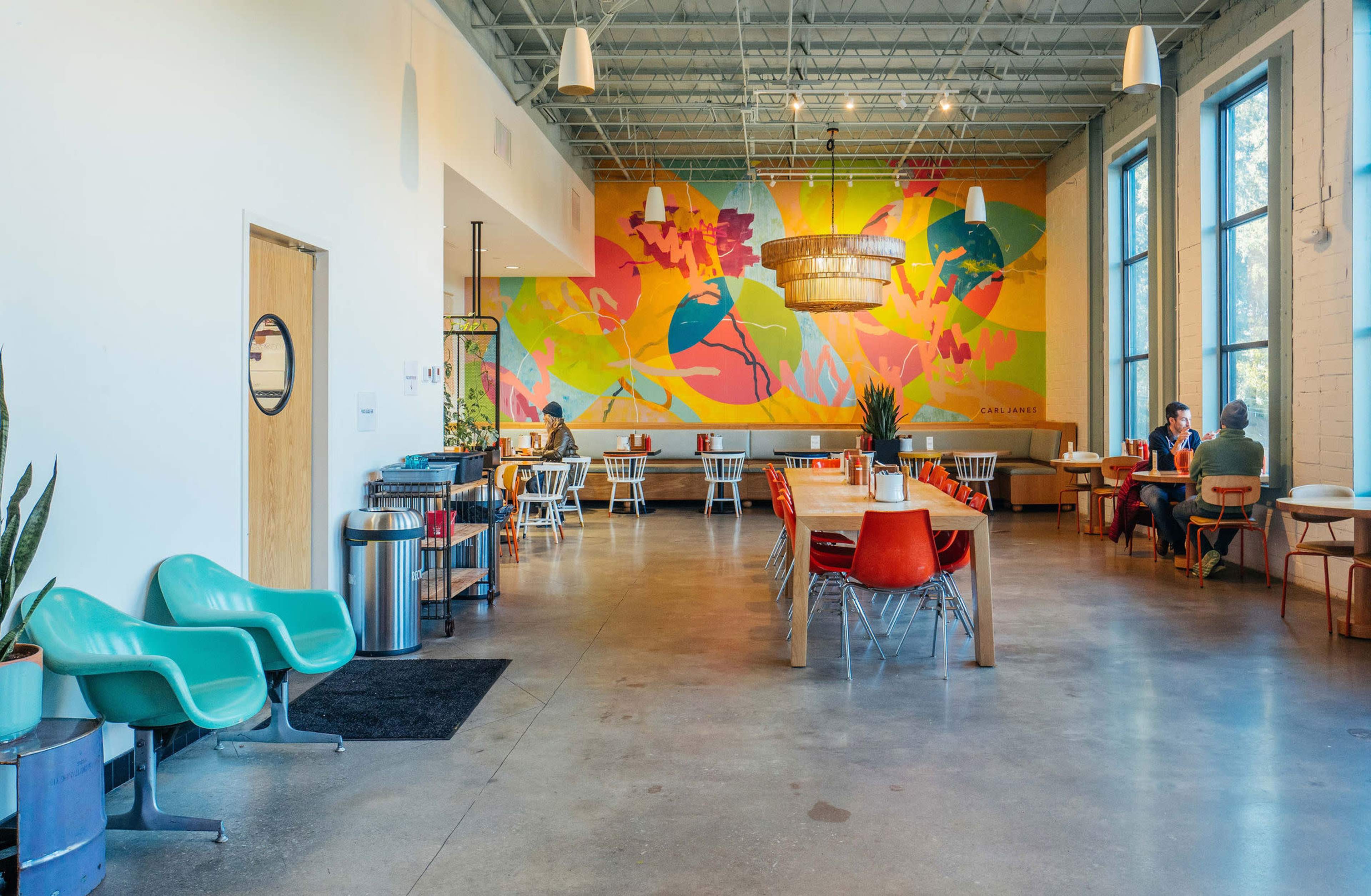 The image shows a spacious, modern interior featuring a vibrant mural, wooden tables, and a mix of seating arrangements in a well-lit café.